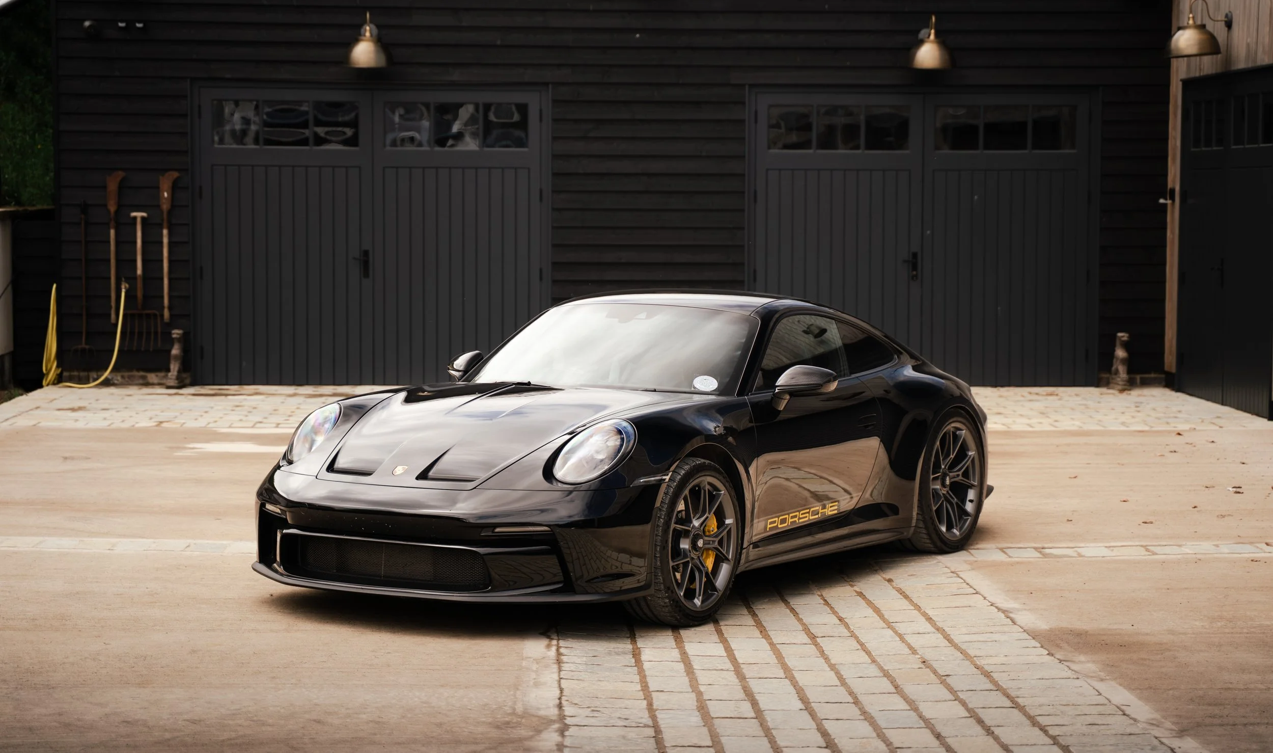 A black Porsche sports car parked in front of a dark wooden garage with 2 garage doors, outdoor lights, and gardening tools on the side.