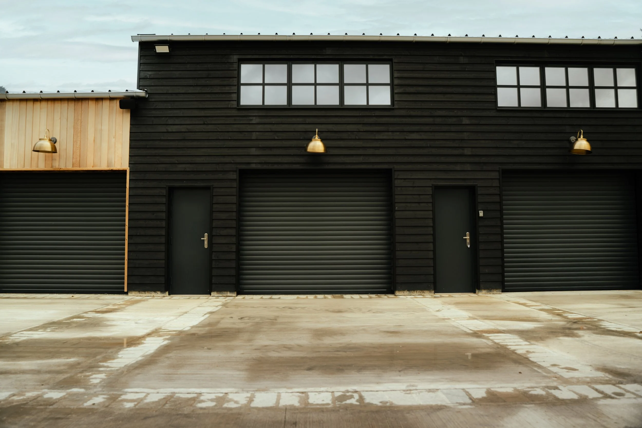 The image features a modern building with a combination of wood and black exterior, including two large garage doors and two smaller doors. There are three golden outdoor wall lights on the building. The ground is concrete with markings, and the sky is cloudy.