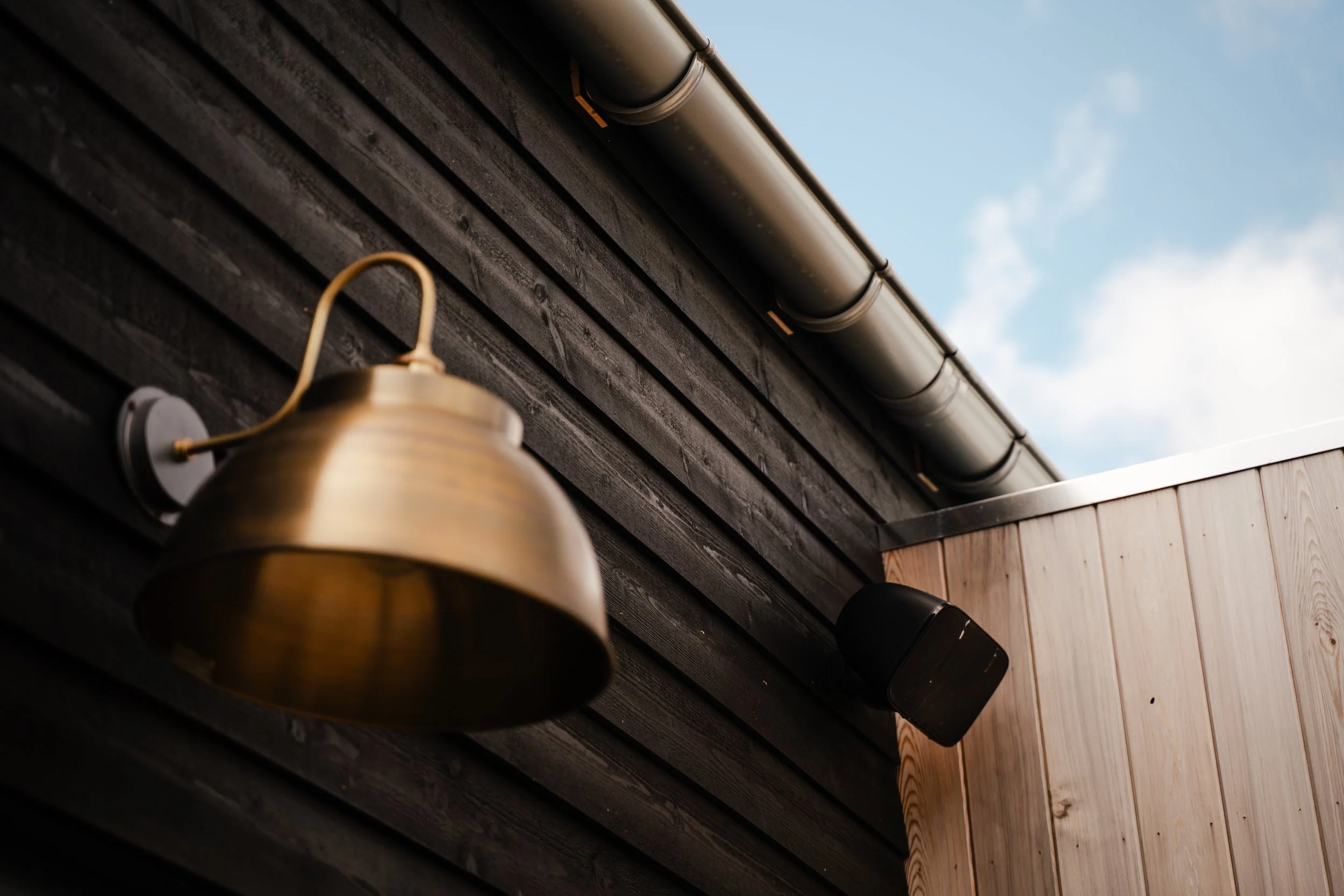 Close-up of a brown house wall with black wooden planks, a gray roof gutter, a brass outdoor light fixture, and a light blue sky with clouds in the background.