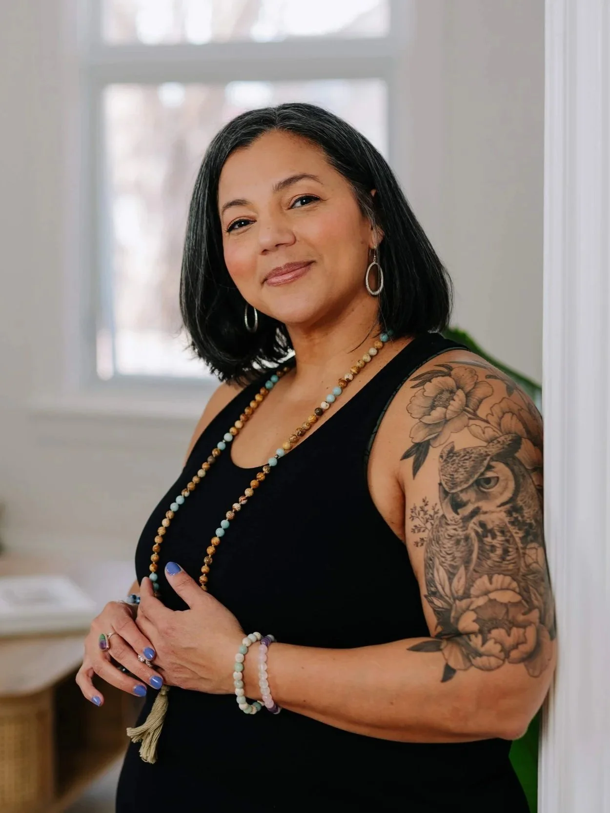A woman with black hair and medium skin tone smiling, wearing a black sleeveless top, colorful beaded necklaces and bracelets, silver hoop earrings, and revealing a tattoo of a bird and flowers on her right arm. She is standing indoors near a white wall and a window.