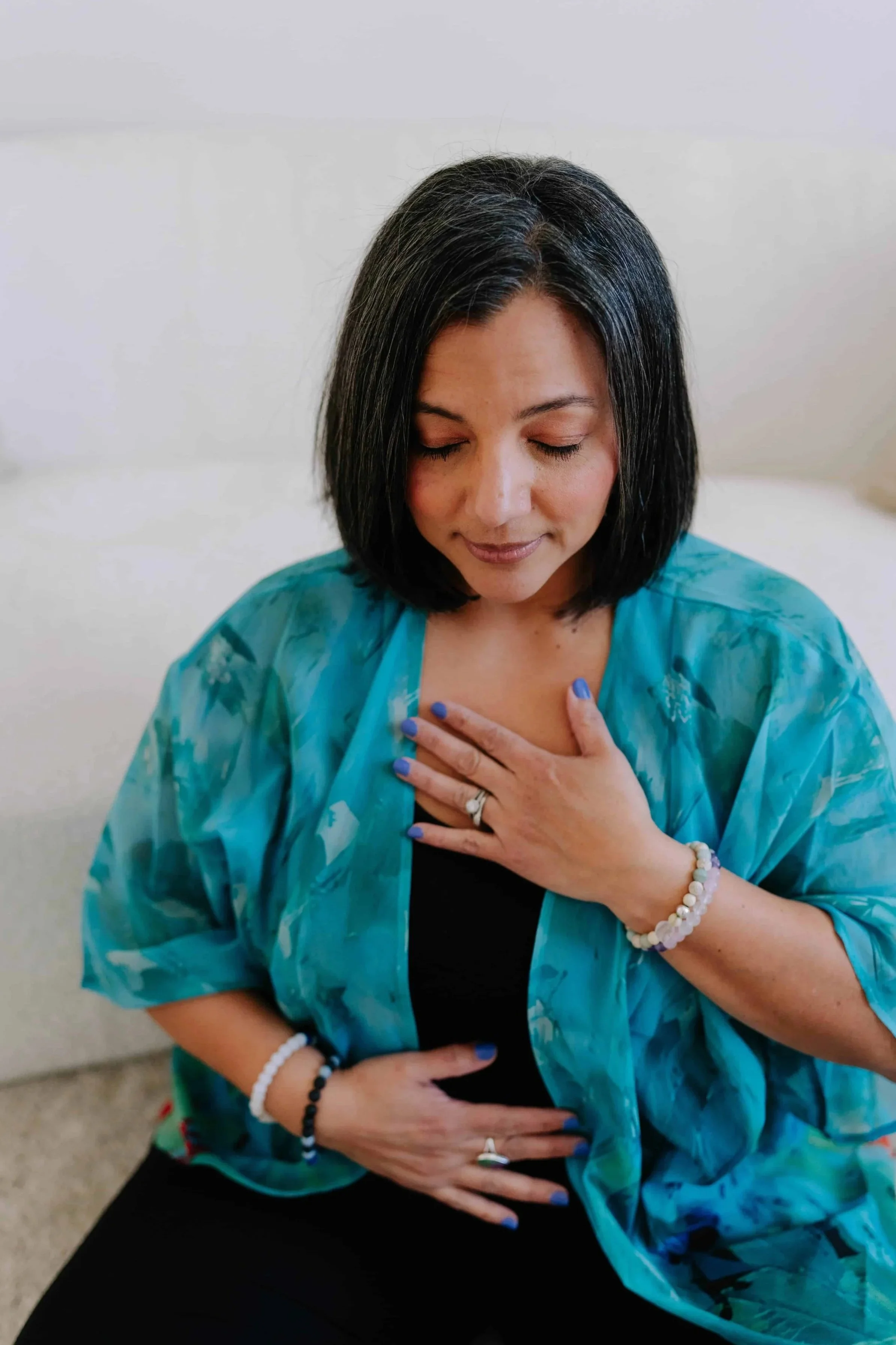 Woman with dark hair and blue flowy shirt with hands on her chest and belly with her eyes peacefully closed.