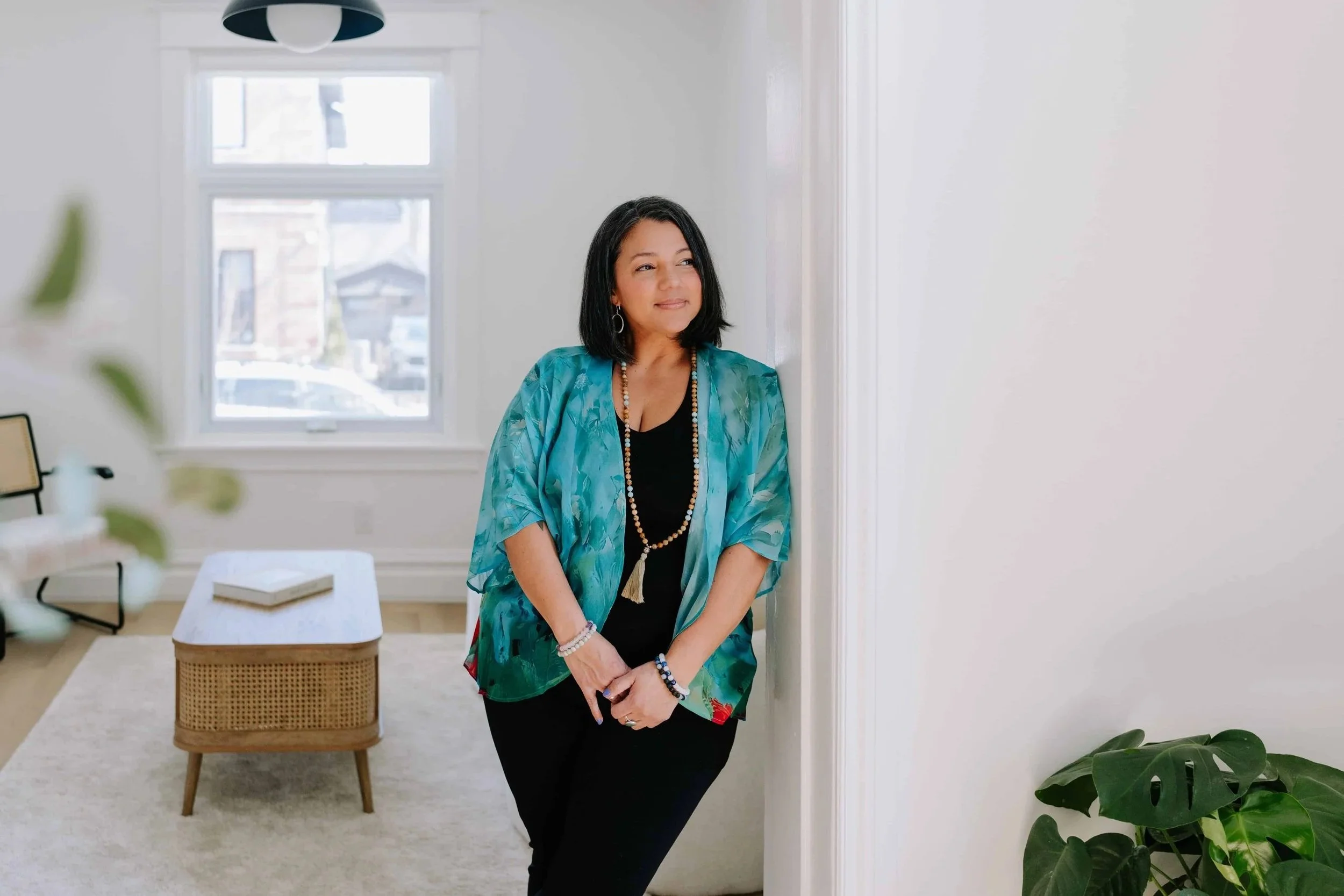 A woman with shoulder-length dark hair wearing a colorful blue-green kimono-style jacket, black top, and black pants, standing in a bright living room next to a white wall, with a window, a coffee table, and a large green plant nearby.