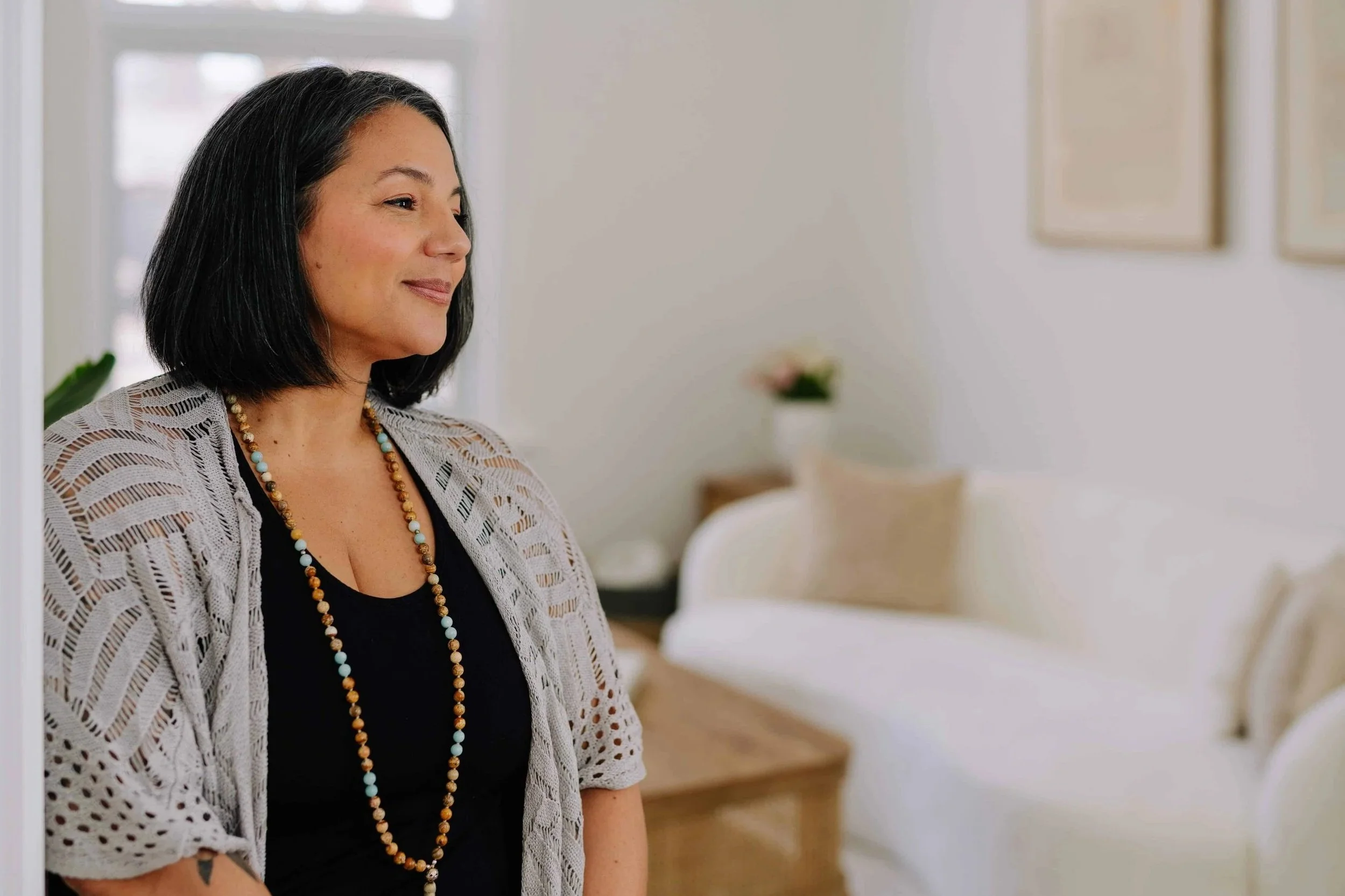 Woman smiling warmly in a white room wearing a black tank top with a beige sweater.