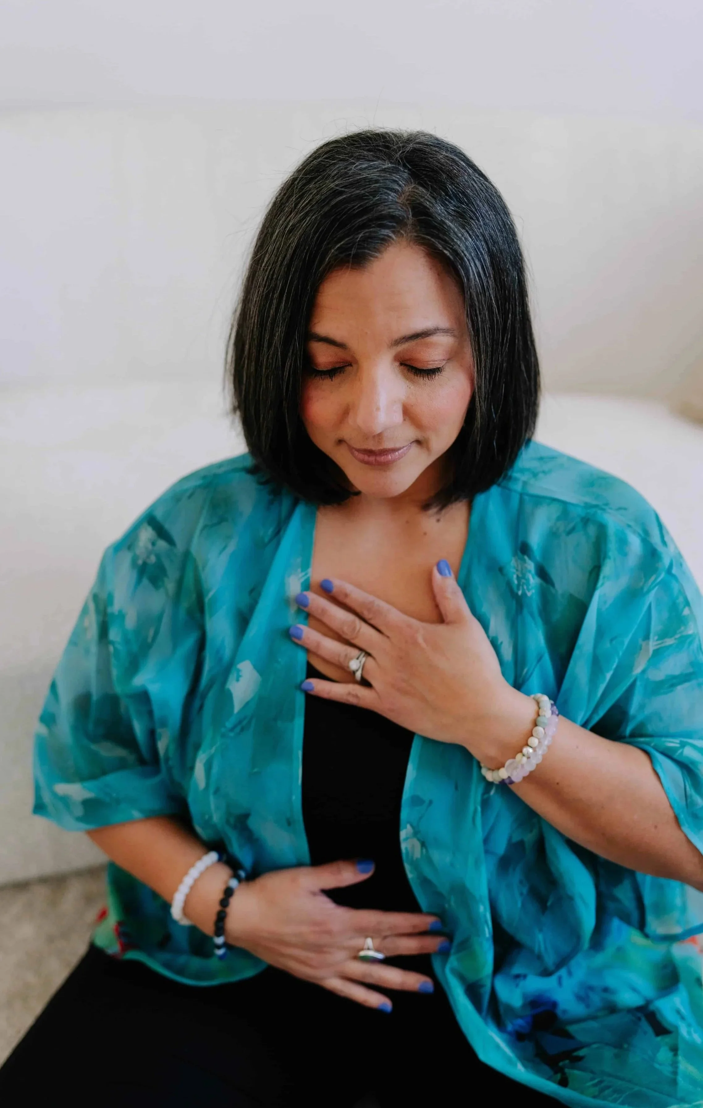 A woman with black hair and blue nails is sitting with her eyes closed, hand on her chest, wearing a blue patterned cover-up and jewelry.