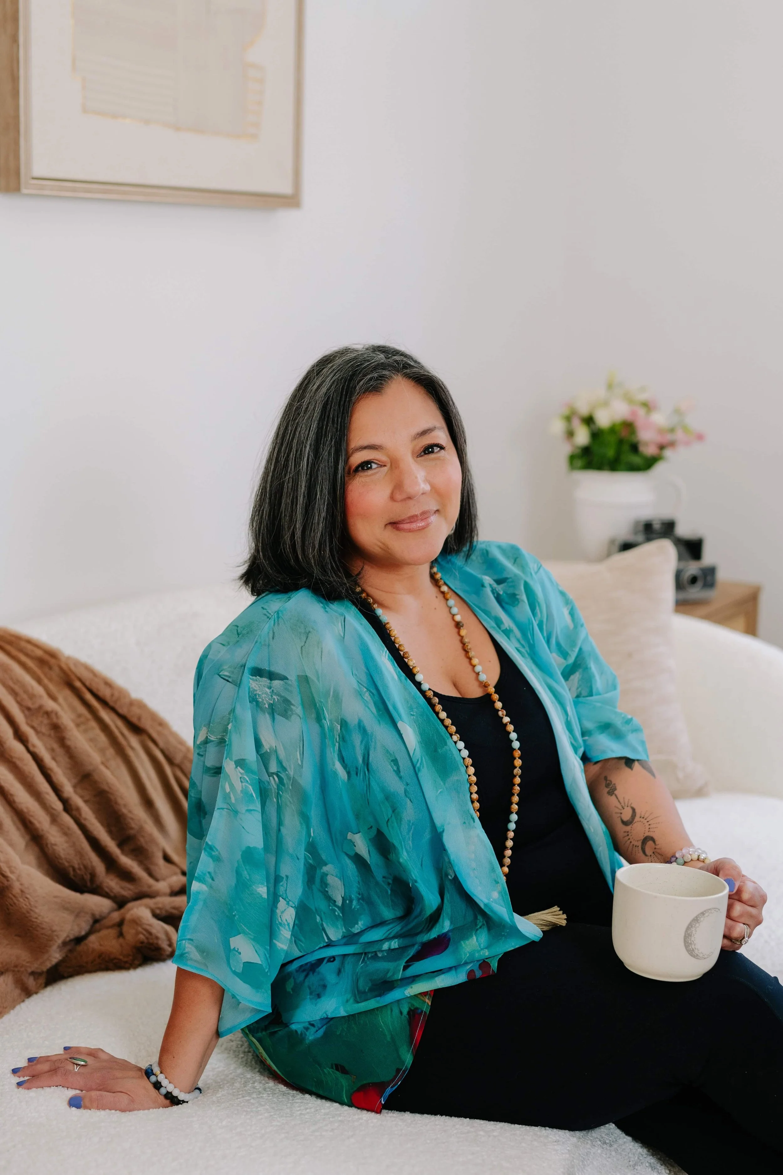 A middle-aged woman with shoulder-length black hair, sitting on a bed with white bedding, holding a white mug with a moon and star design. She is wearing a black top, a turquoise patterned cover-up, and beaded jewelry. She has tattoos on her right arm and is smiling softly. Behind her, there are pillows, a nightstand with a potted plant with pink and white flowers, and a camera.