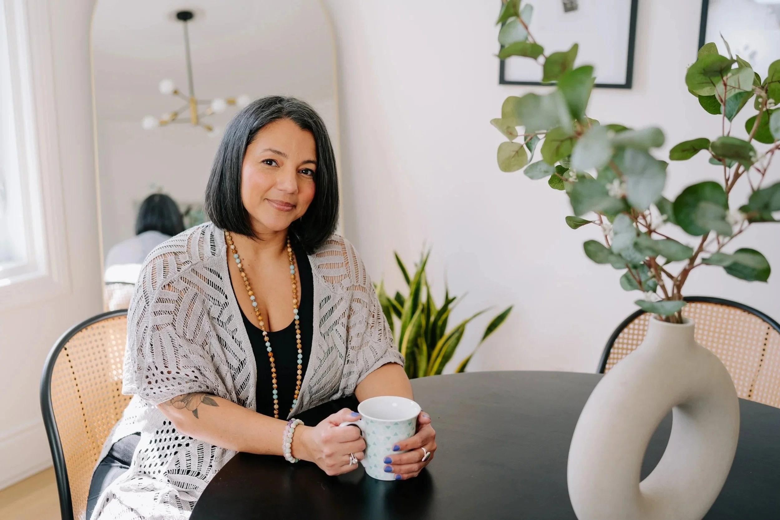 A woman with shoulder-length black hair and a tattoo on her left arm, sitting at a table with a cup of coffee, in a well-lit room with a plant in a decorative vase and framed artwork on the wall.
