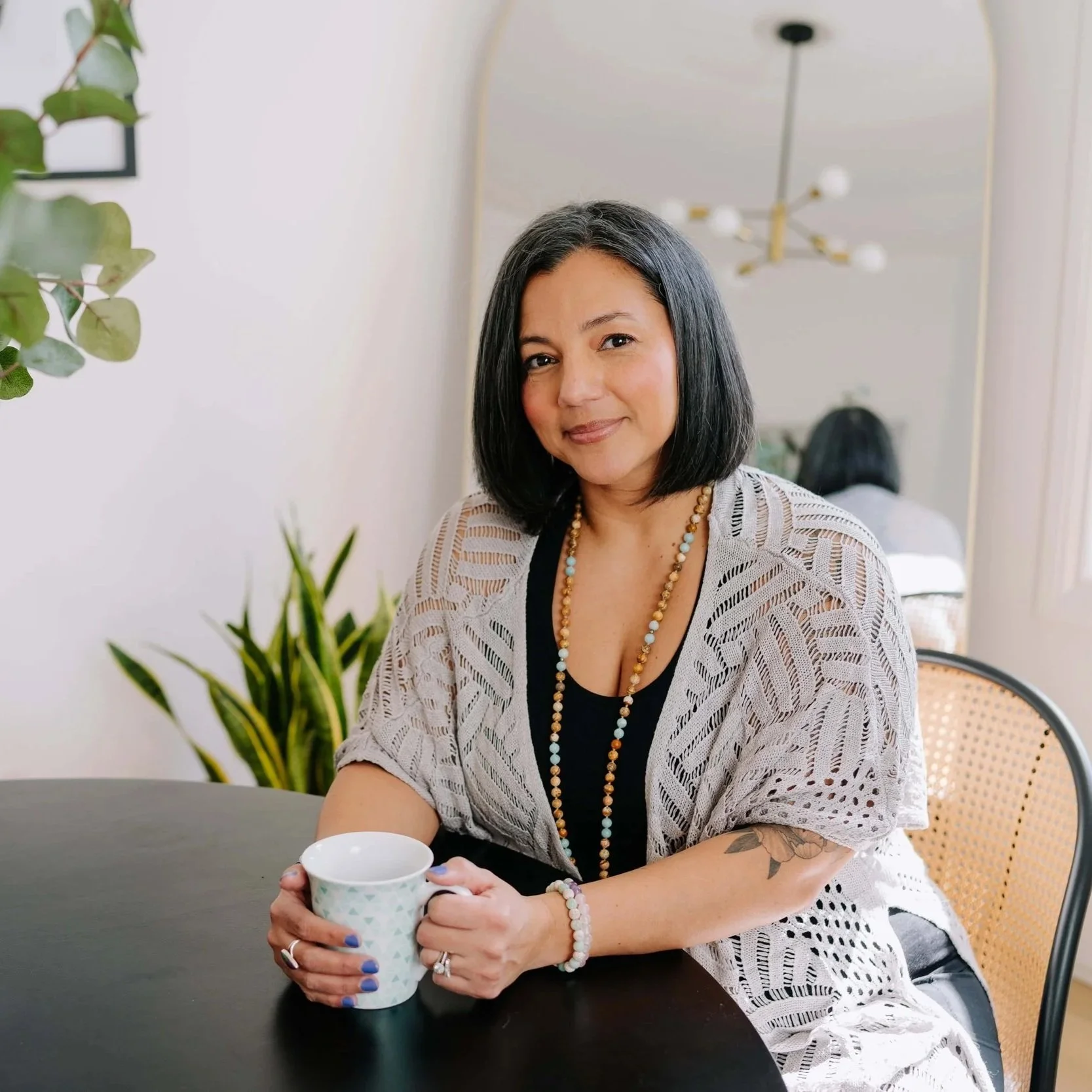 Woman holding tea cup with beige sweater sitting at table gently smiling.