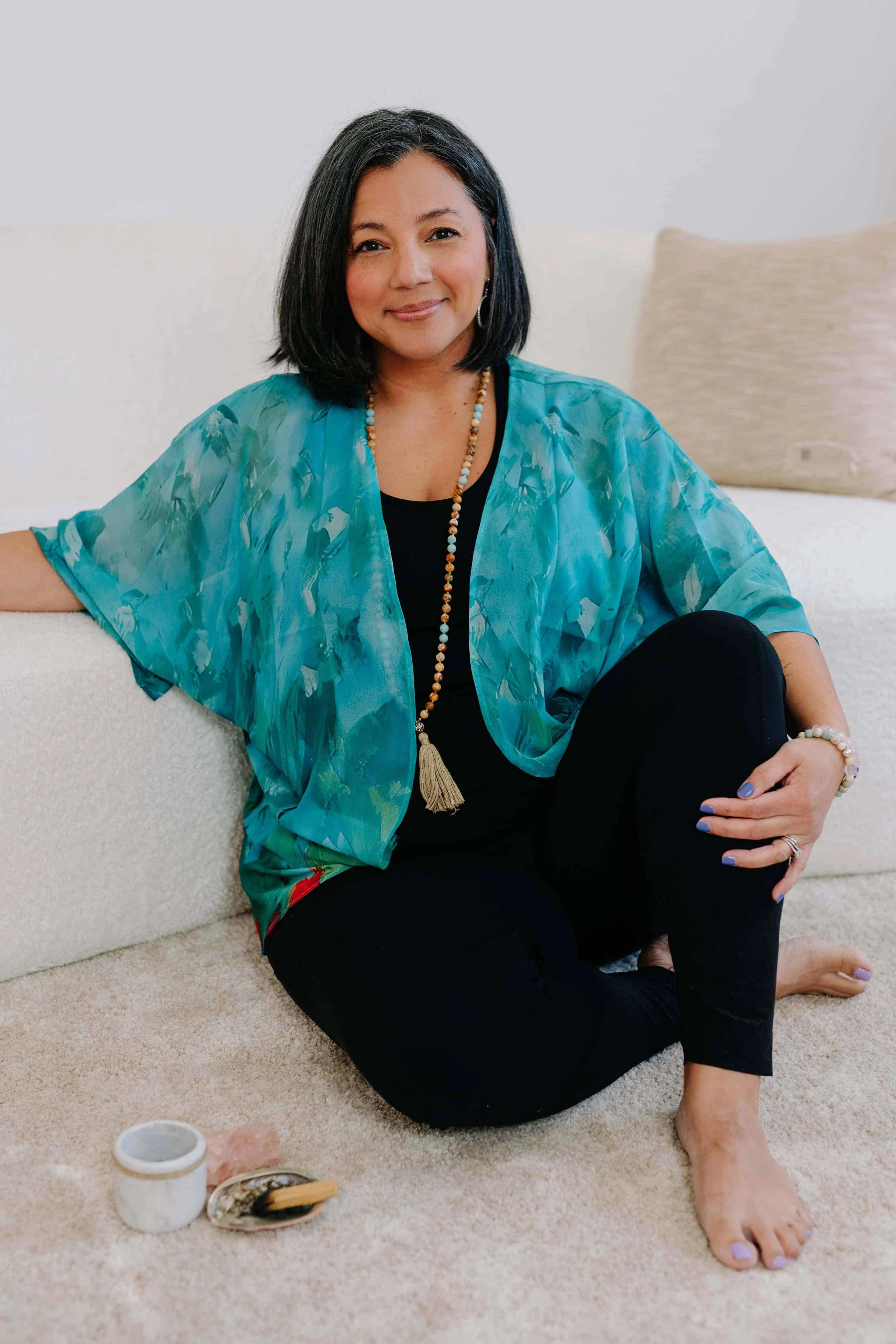 A woman sitting on a beige carpeted floor, leaning against a white sofa. She is wearing a turquoise kimono-style top, black pants, and jewelry including a necklace, bracelet, and ring. She has shoulder-length black hair and is smiling at the camera. There are small objects including a candle and a decorative box on the floor in front of her.