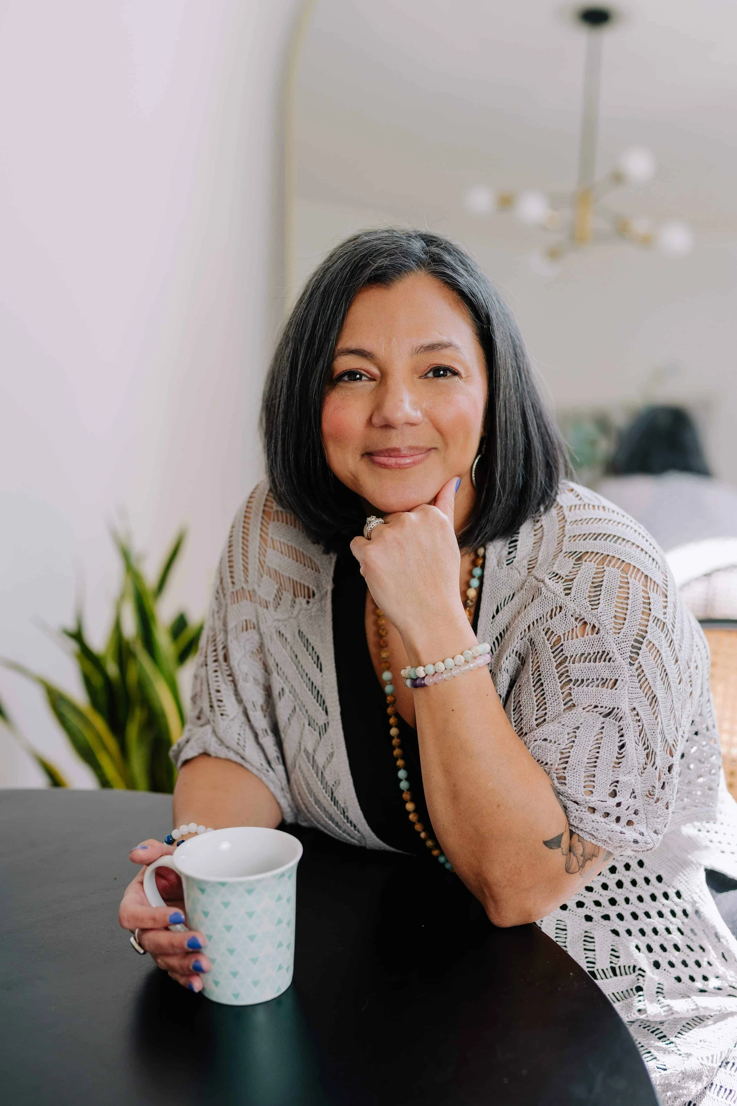 Woman in beige sweater, wearing long beads on neck and holding a teal tea cup smiling with her hand on her chin.