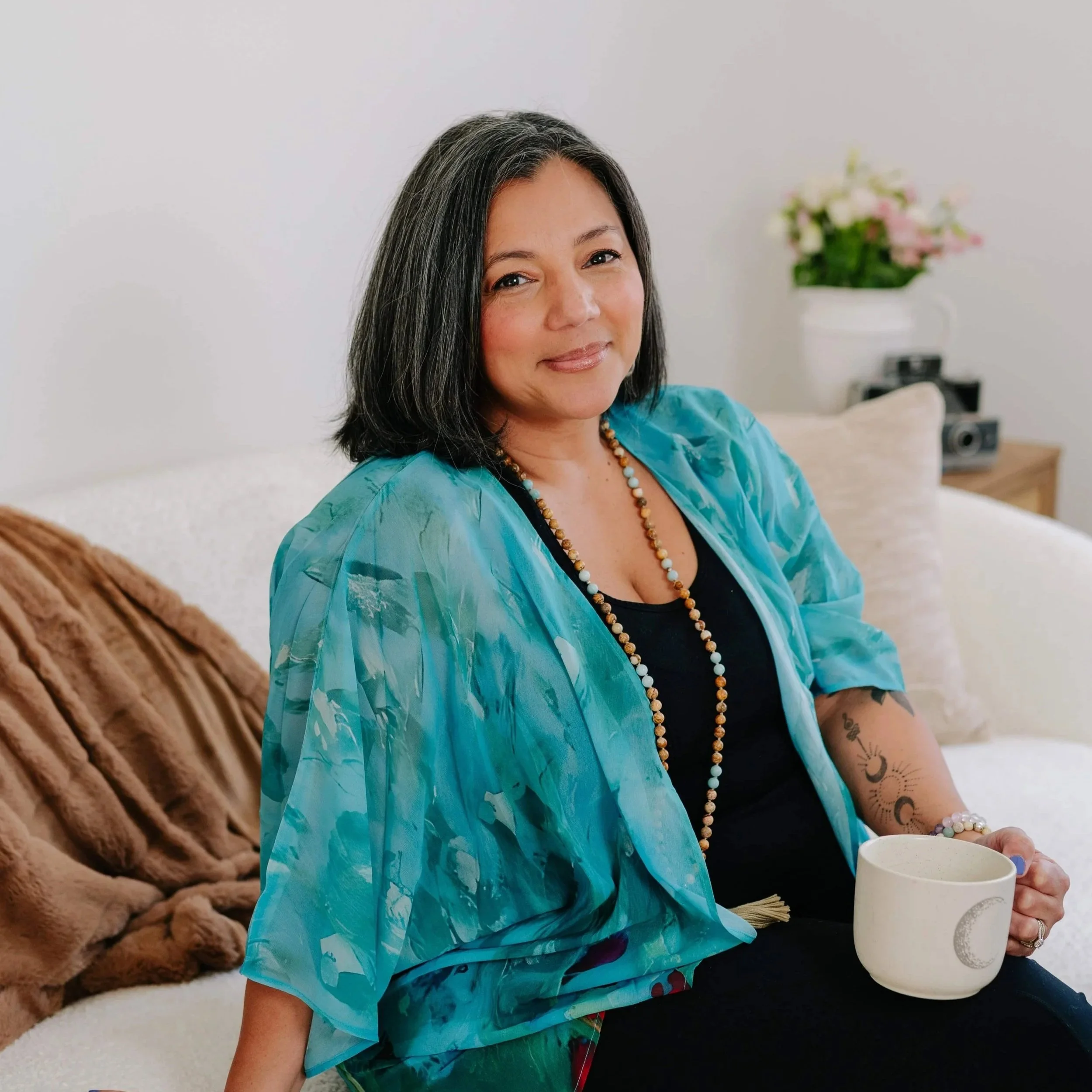 A woman with shoulder-length black hair, wearing a turquoise shawl, black top, and beaded necklaces, sits on a white sofa holding a white mug with a moon design. In the background, there is a flower vase and camera on a side table.