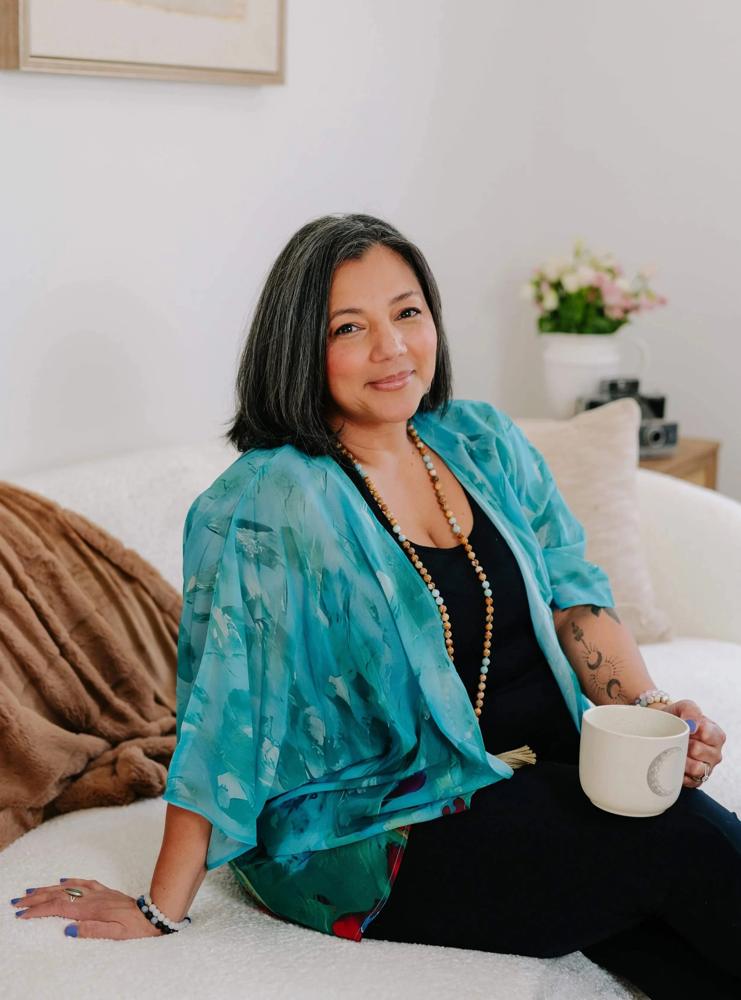 A woman with medium-length black hair, wearing a colorful blue paisley shawl and black clothing, sitting on a white bed holding a white mug with a moon and star design. She is smiling softly, with tattoos on her right arm, accessorized with a beaded necklace and bracelets. The background includes a beige blanket, a dresser with a flower vase, and a camera on the side.