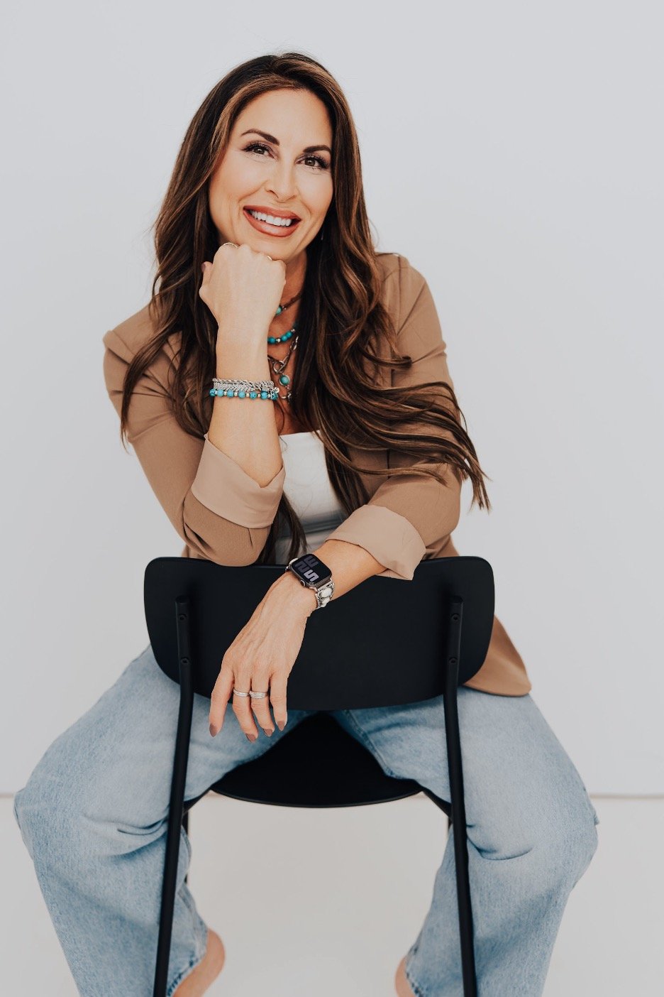 A smiling woman with long brown hair, wearing a tan blazer, white top, and light blue jeans, sitting on a black chair against a plain white background.