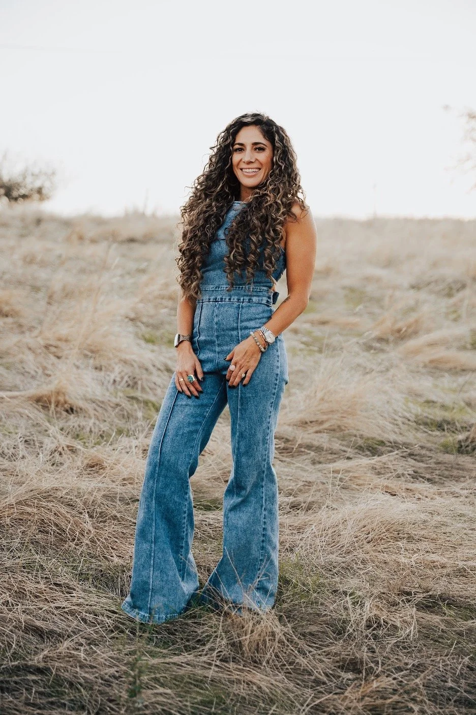 A woman standing in a grassy field, smiling, wearing a sleeveless denim jumpsuit with wide-leg pants and accessories.