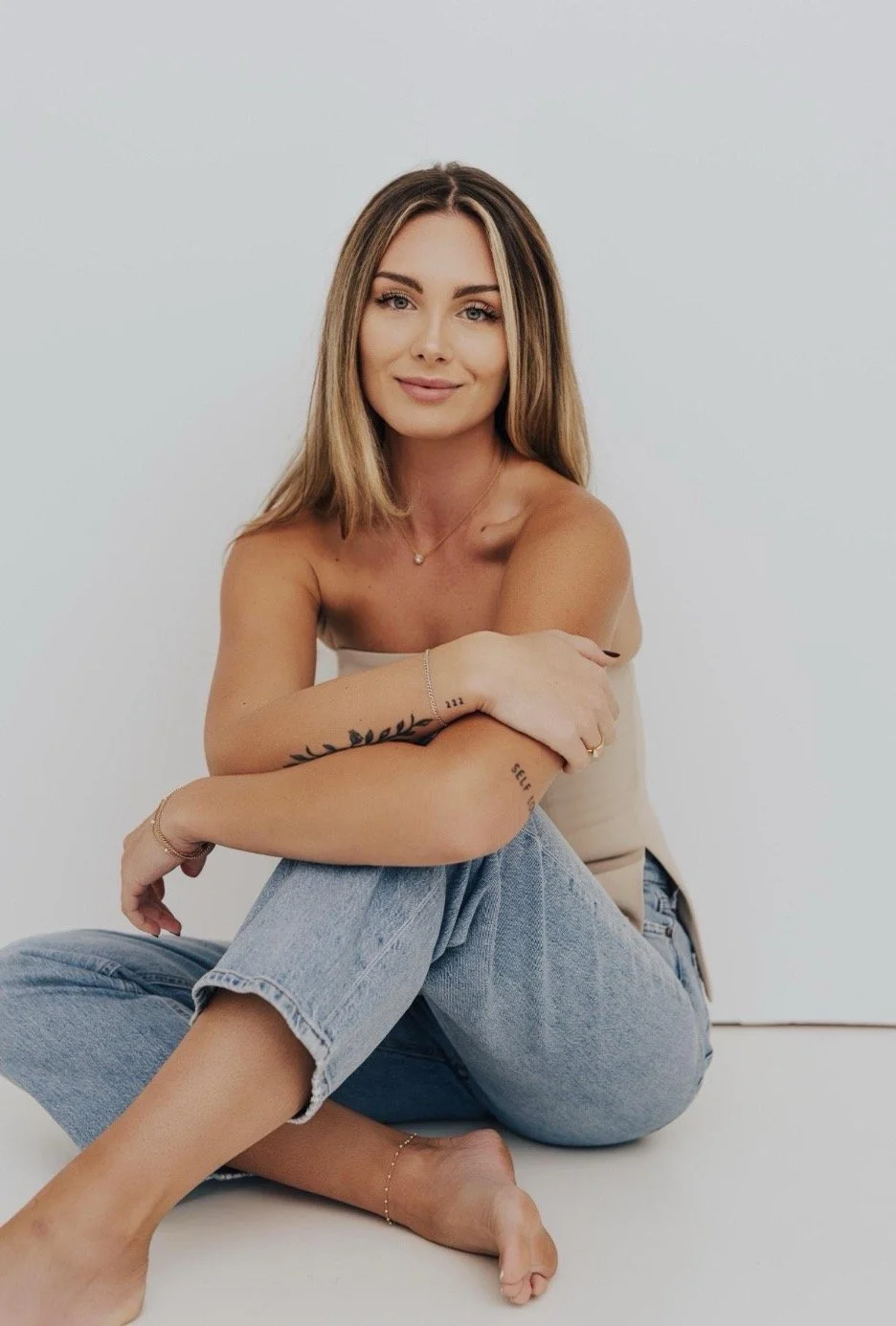 A young woman with long light brown hair, sitting cross-legged against a plain white background. She is smiling gently, with her arms crossed over her knees, wearing a beige strapless top and light blue jeans, with a delicate necklace and bracelets on her wrist.