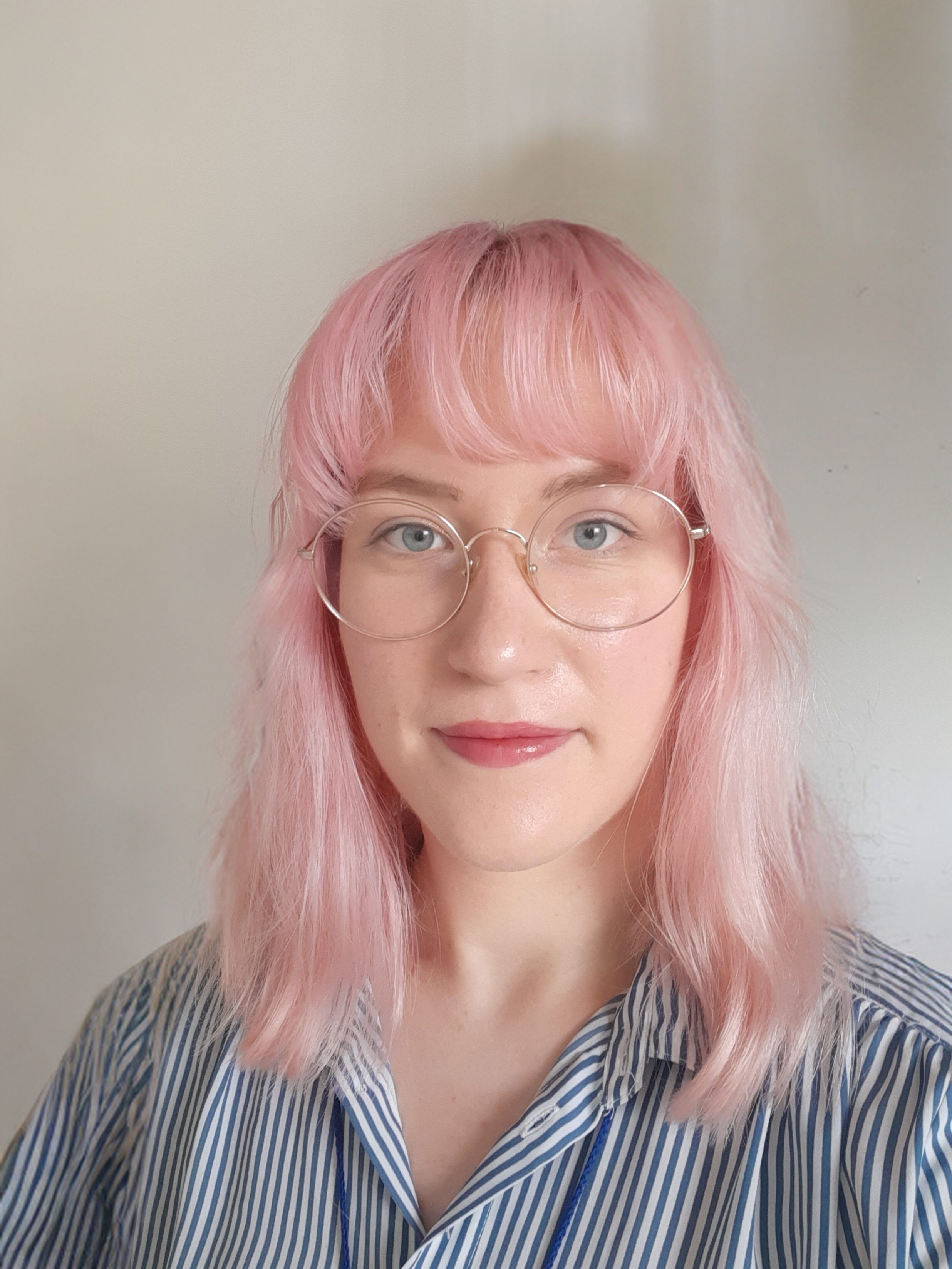 A woman with light pink hair, round glasses, and a striped shirt posing in front of a plain background.
