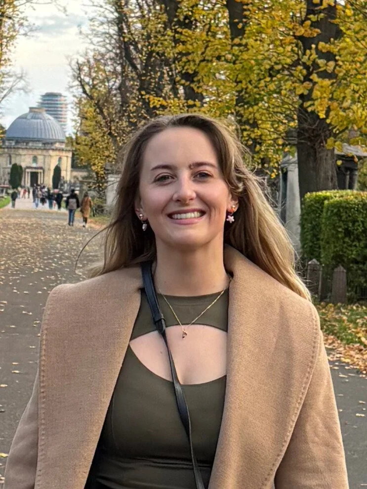 A young woman with light brown hair smiling outdoors in a park during autumn. She is wearing a beige coat over an olive green top, with trees in the background.