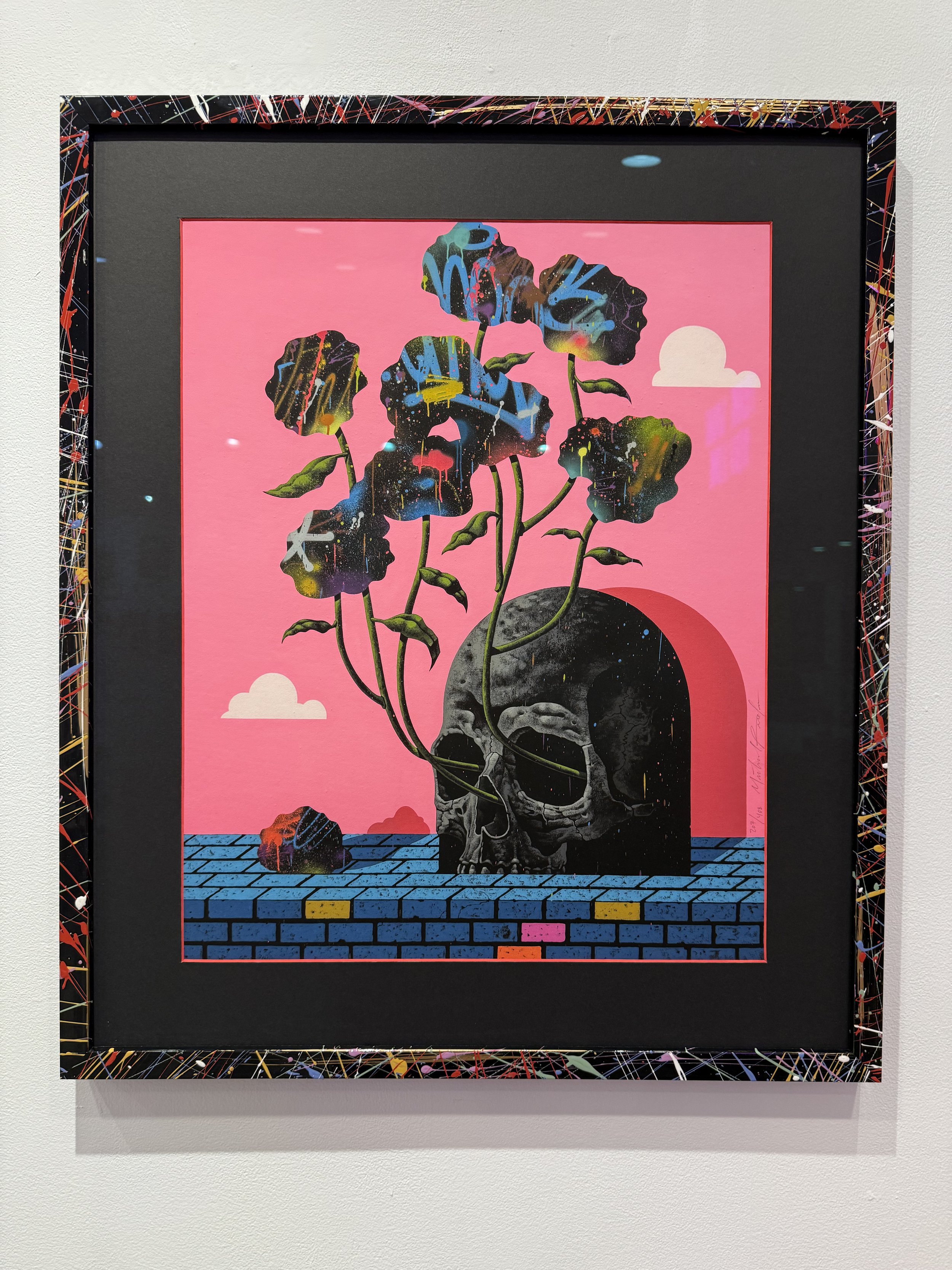 A vibrant artwork featuring a black skull with large openings for eyes and nose, with colorful plants growing from it against a pink sky with clouds. The background includes a blue brick platform with some multicolored bricks and a pink rock. The art