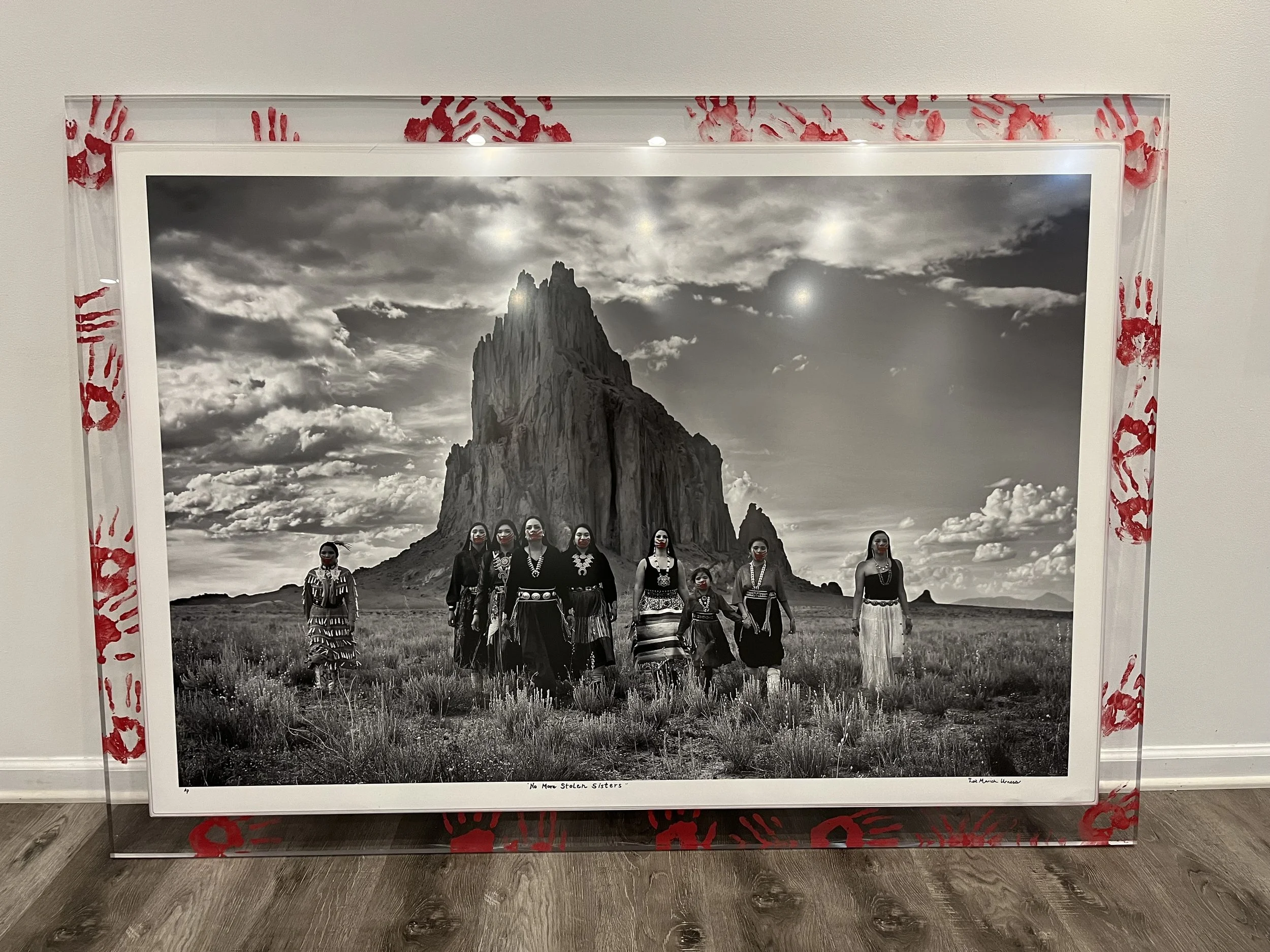 Black and white photograph of nine women wearing traditional clothing, standing in a grassy field with a large mountain in the background, surrounded by a frame decorated with red handprints.