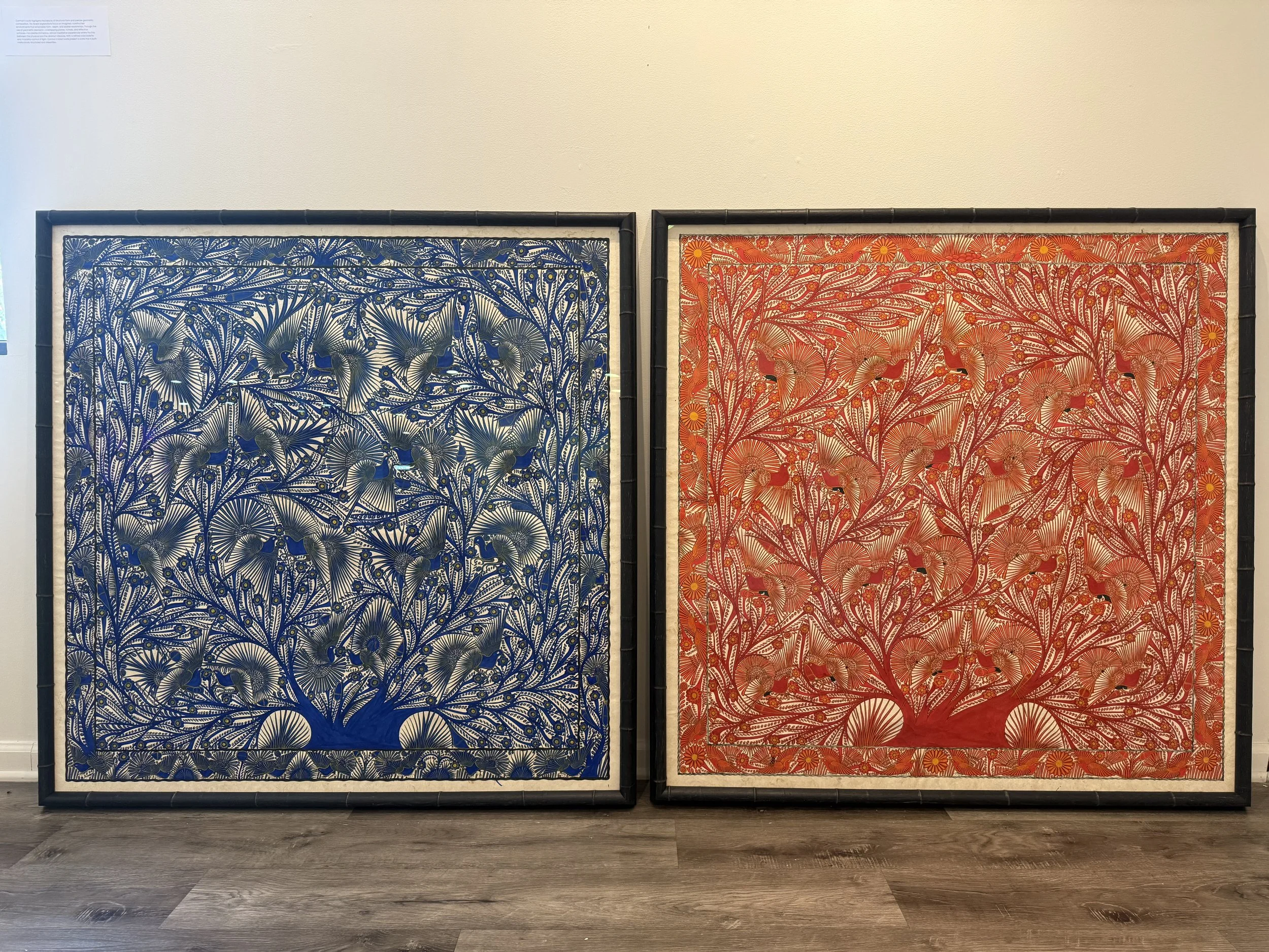 Two framed textile artworks with elaborate floral and bird patterns, one blue and the other red, leaning against a plain white wall.