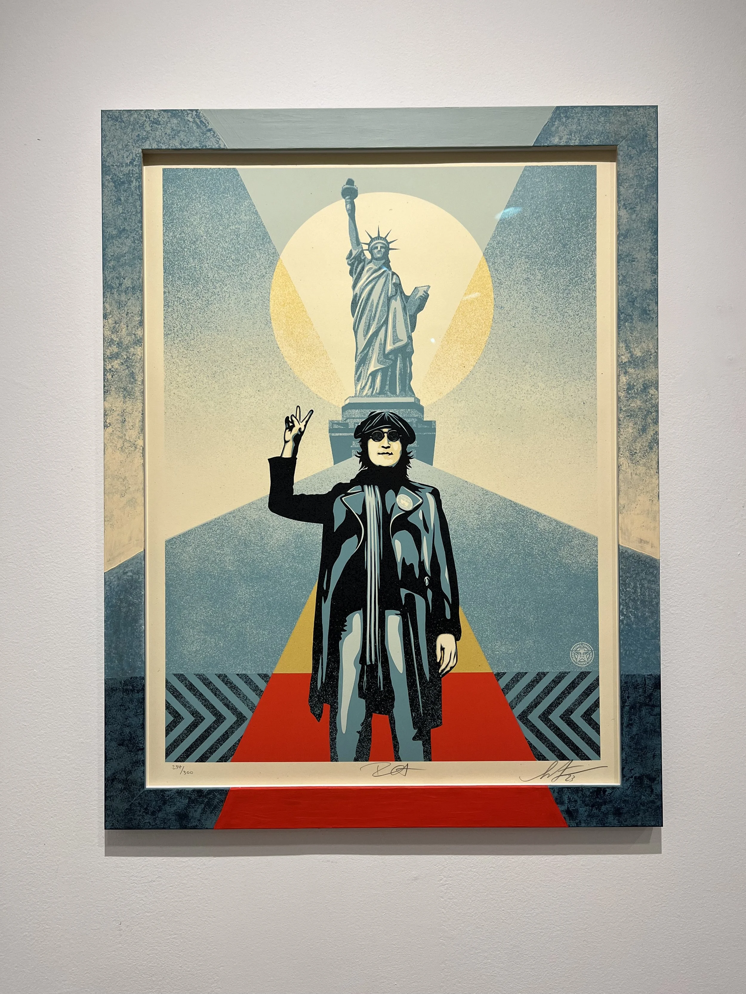 A modern graphic art print depicting a person in black clothing and sunglasses standing on a red carpet, raising one hand in a peace sign in front of the Statue of Liberty.