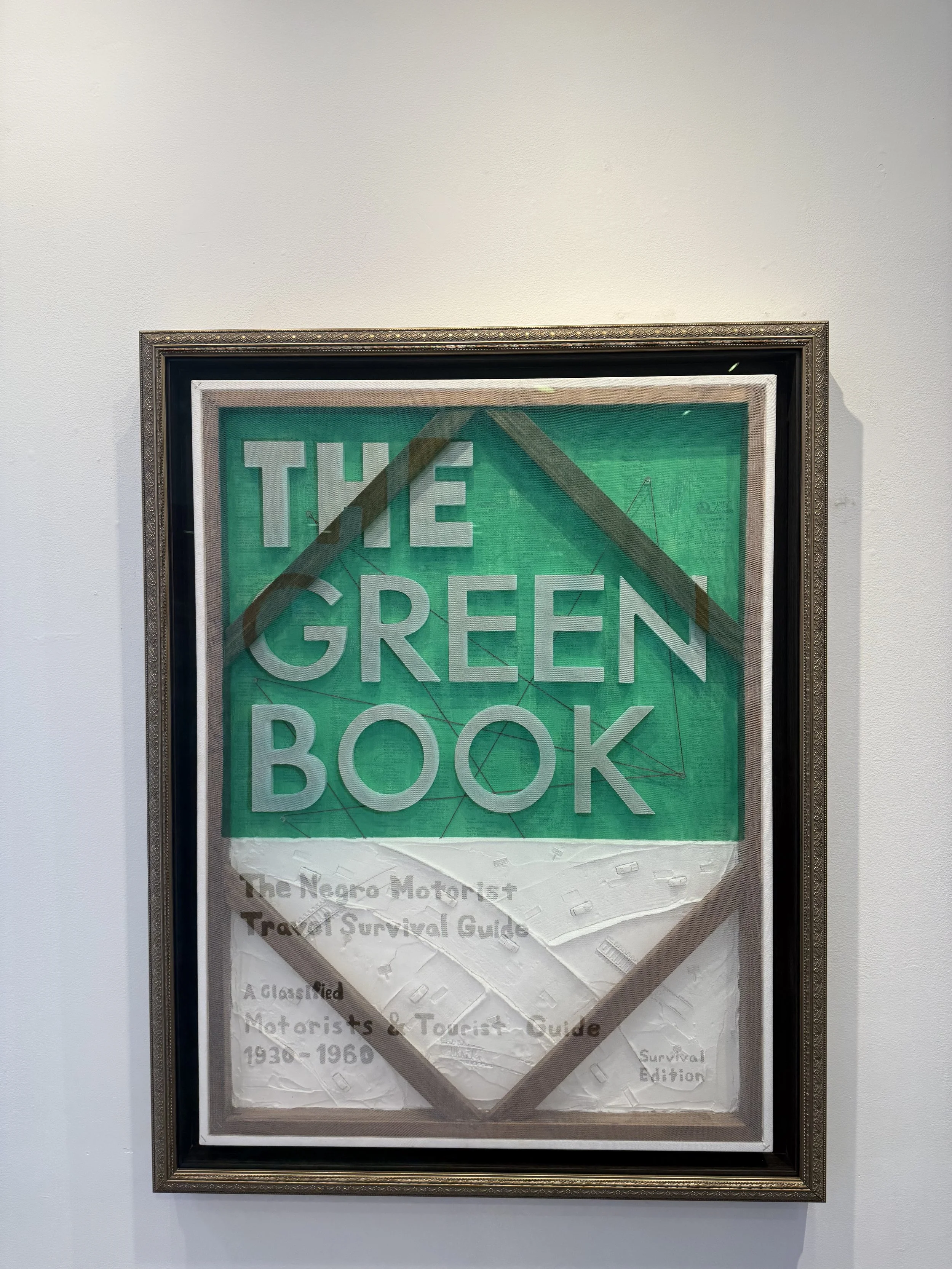 Framed art piece titled 'The Green Book,' featuring a green background with large gray text and a white and brown geometric design. Smaller text at the bottom reads 'The Negro Motorist Travel Survival Guide' and 'A Classified Motorists & Tourist Guid