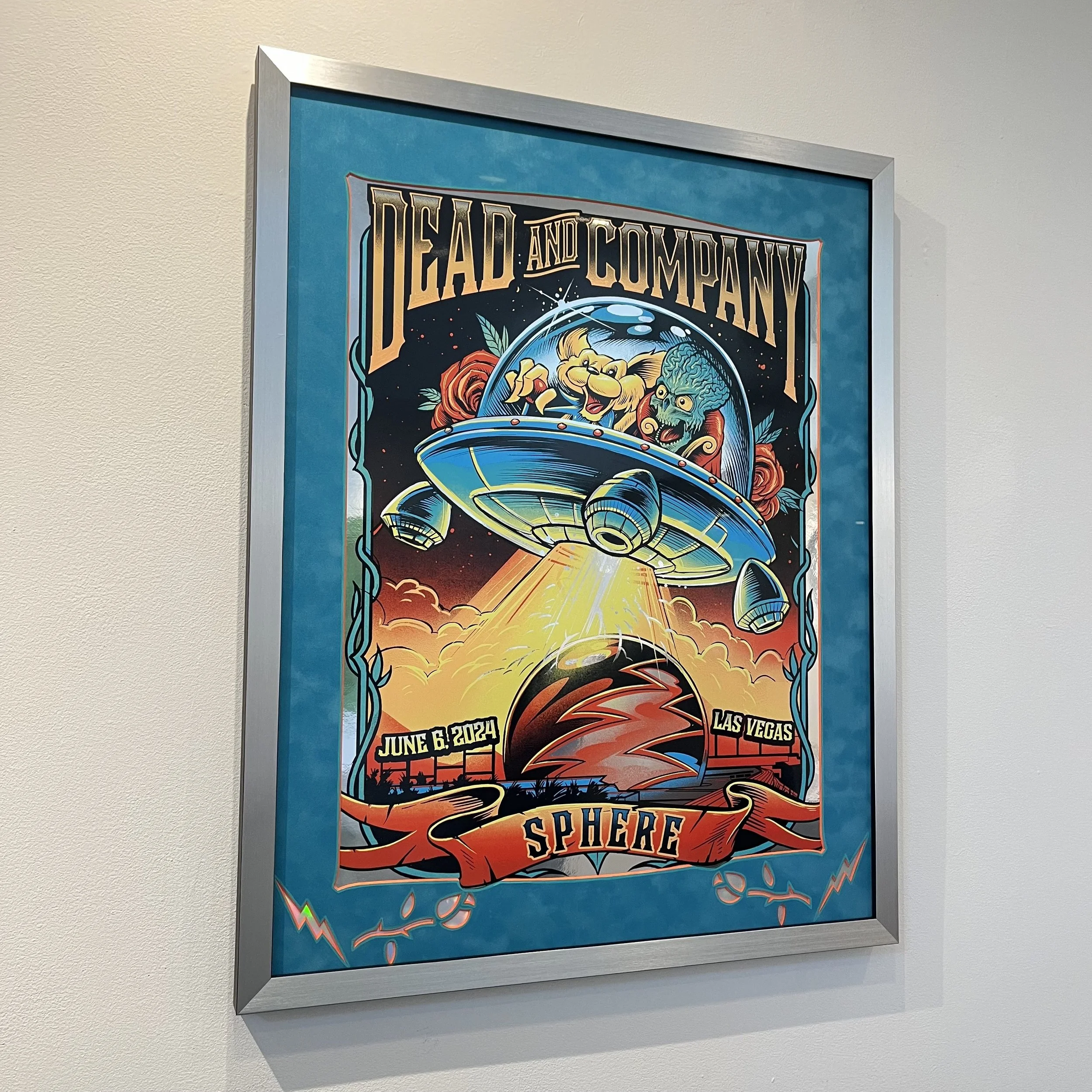 Colorful poster advertising a Dead and Company concert on June 6, 2024, in Las Vegas at Sphere, featuring a spaceship with cartoon characters inside flying over a landscape with a large sphere and sunset background.