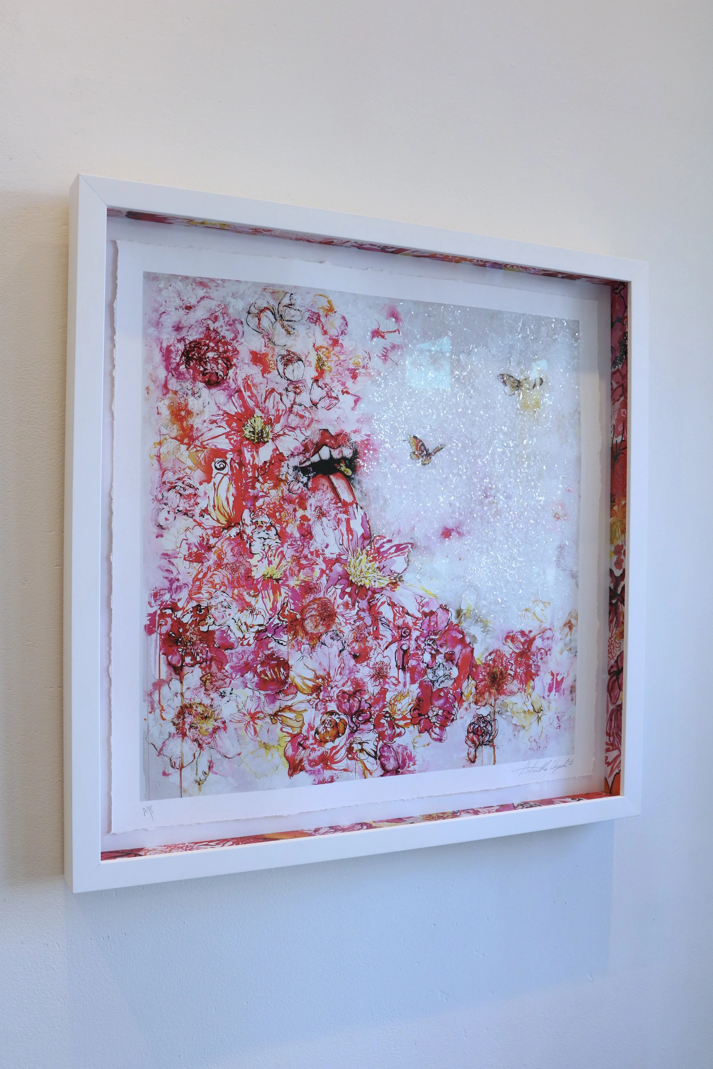 A white-framed artwork hanging on a beige wall features an abstract portrait of a woman's face with her tongue sticking out, surrounded by vibrant red, pink, yellow, and orange flowers, with butterflies and a glittery background.