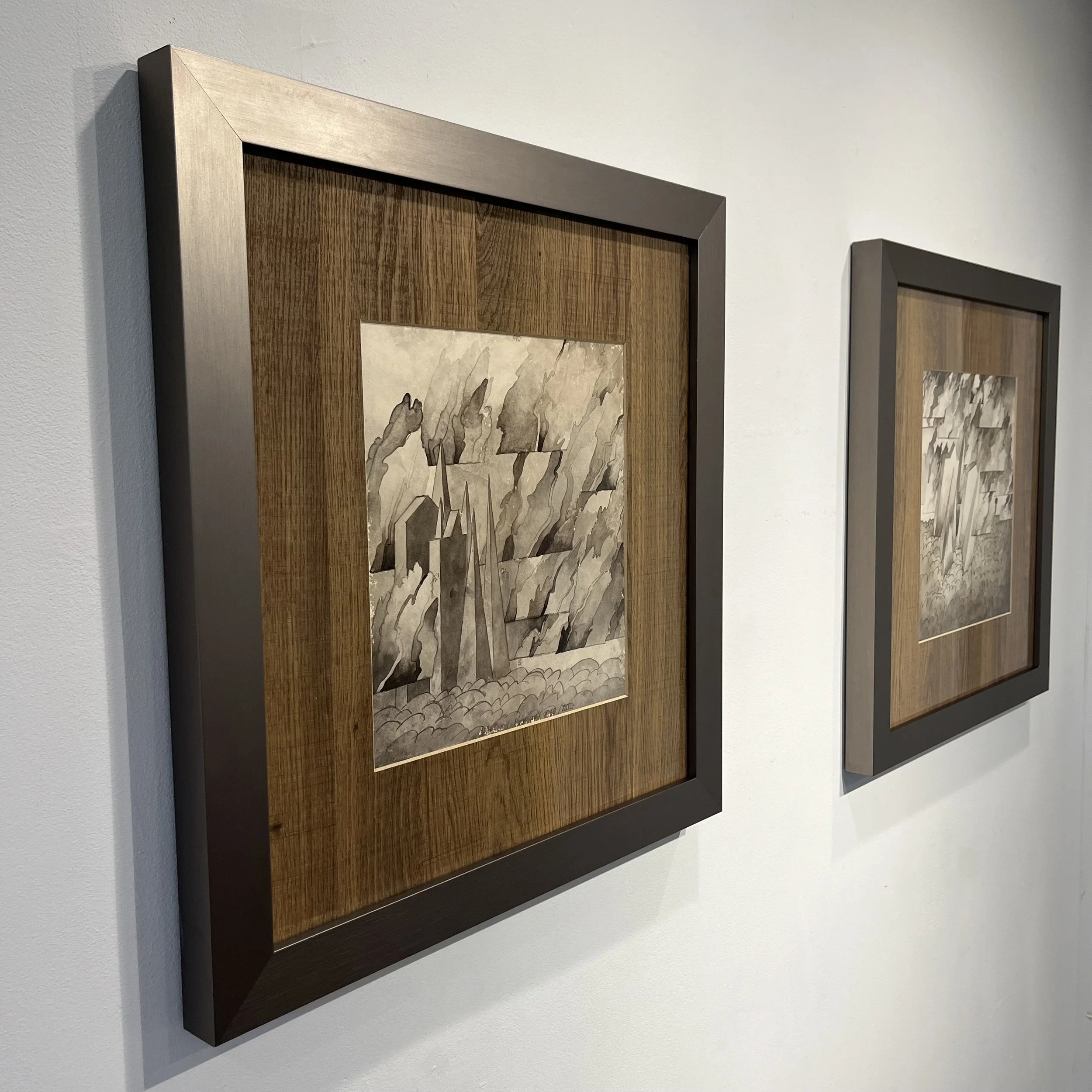 Two framed monochrome landscape paintings hanging on a white gallery wall