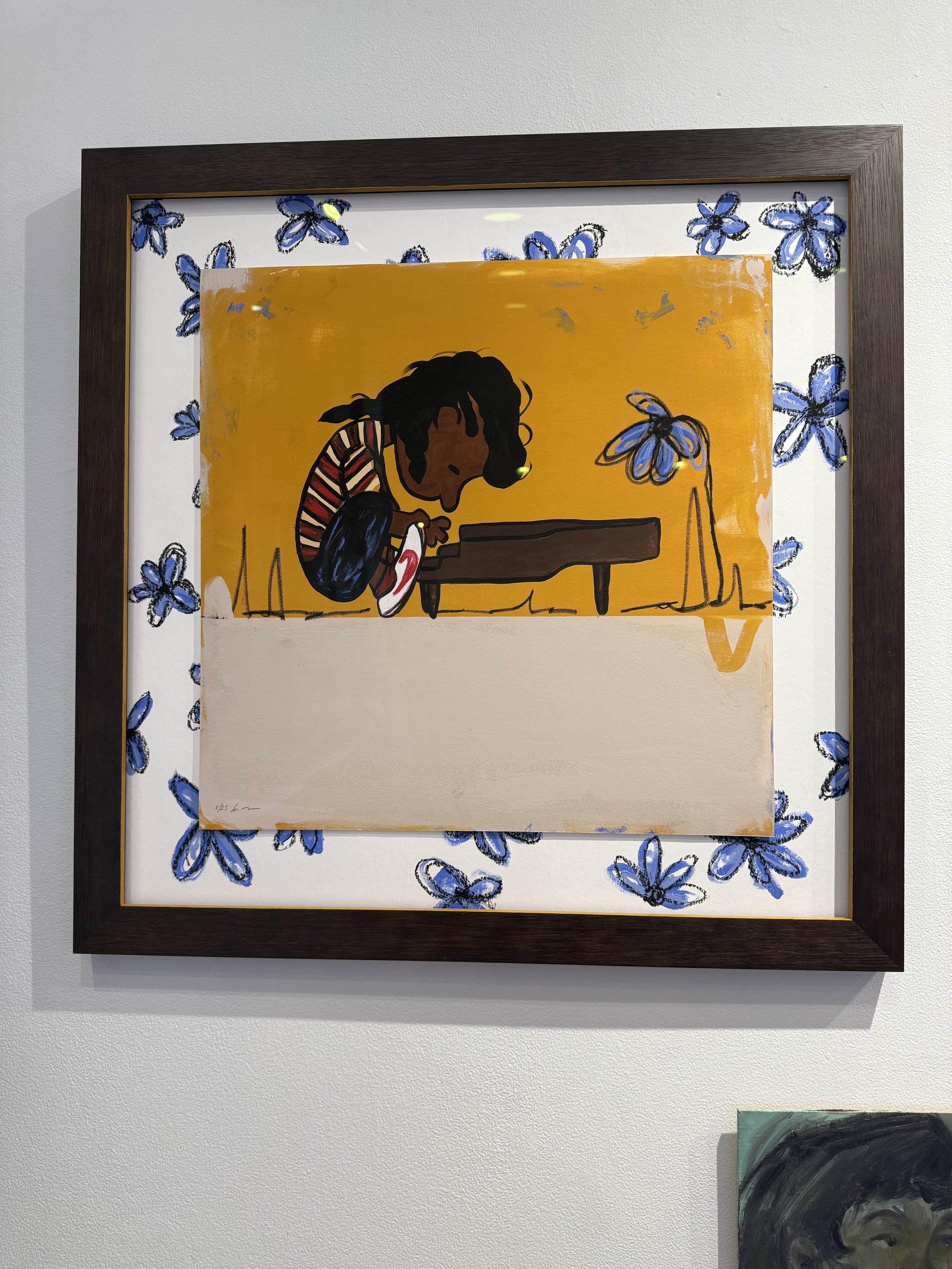 A colorful illustration of a child with dark skin and black curly hair, wearing a striped red and white shirt, sitting on the ground and playing a brown piano against an orange background. The image is surrounded by a white border with blue flowers, and the artwork is framed with a dark brown wooden frame.