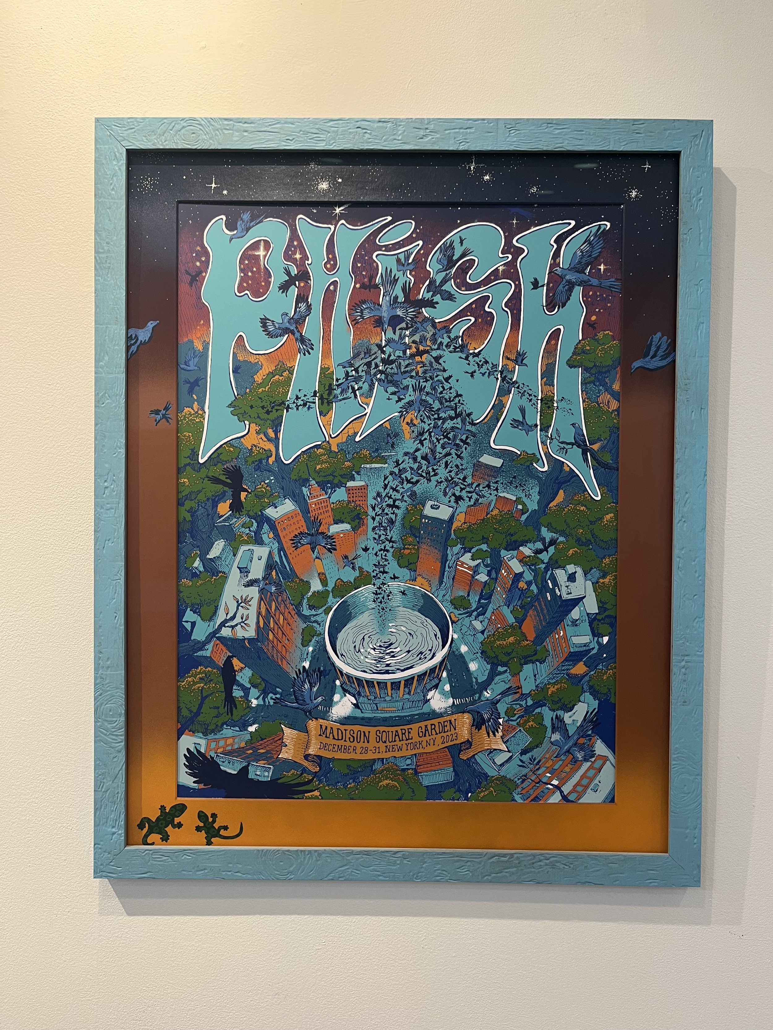 Colorful poster advertising Madison Square Garden from December 28-31, 2023, featuring a cityscape with buildings, trees, many flying birds, and a large water fountain in the center, framed in a blue frame with two small lizard illustrations at the b