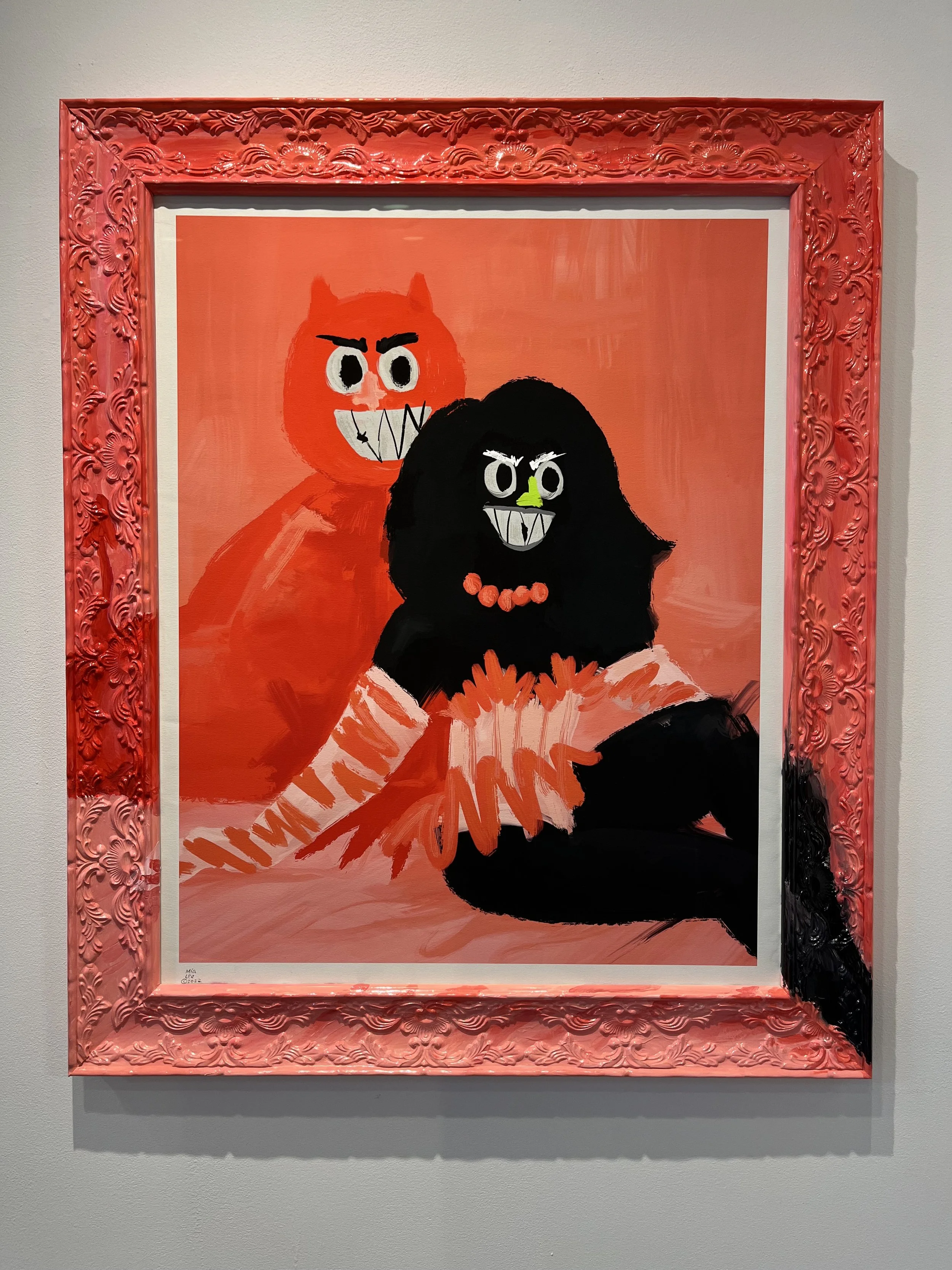 An abstract painting of two monsters with exaggerated features, one in red and one in black, with a pink ornate frame, displayed on a gray wall.