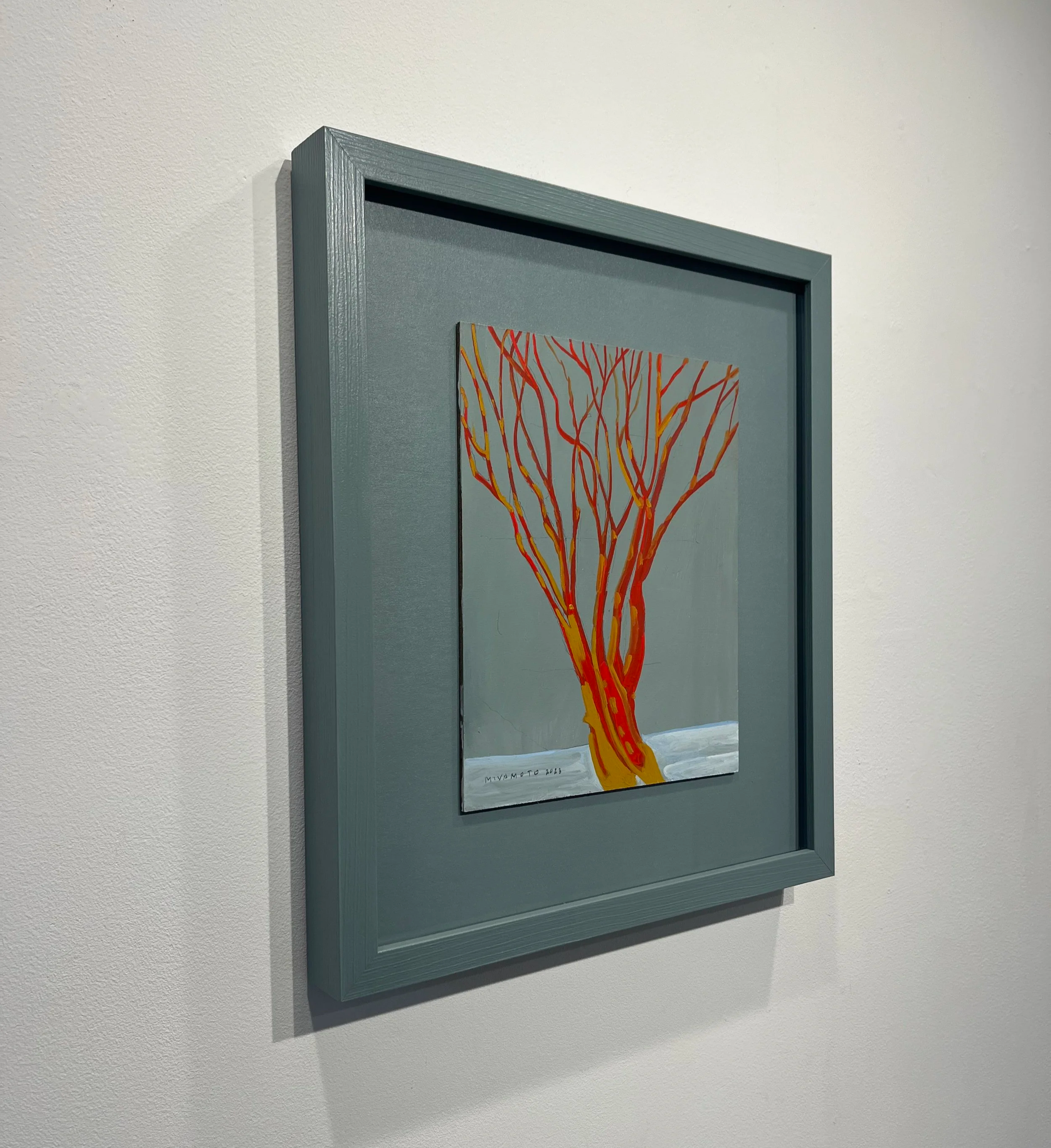 Framed artwork featuring a colorful abstract tree with orange, red, and yellow branches set against a light background, hung on a white textured wall.