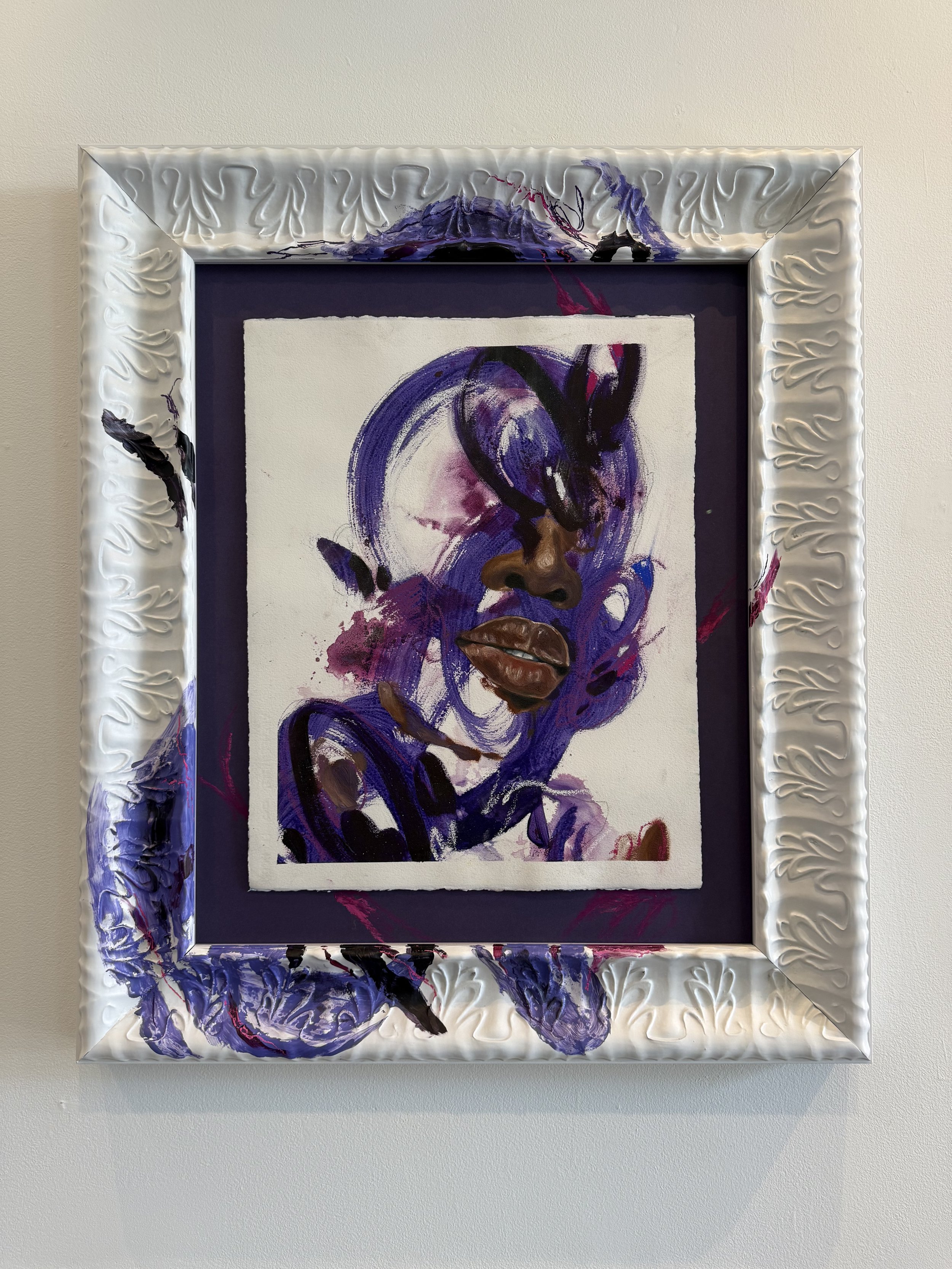 A mixed media artwork of a woman's face with dark brown lips, painted with purple, black, and brown paint on paper, set in an ornate white frame with purple, black, and red paint splashes.