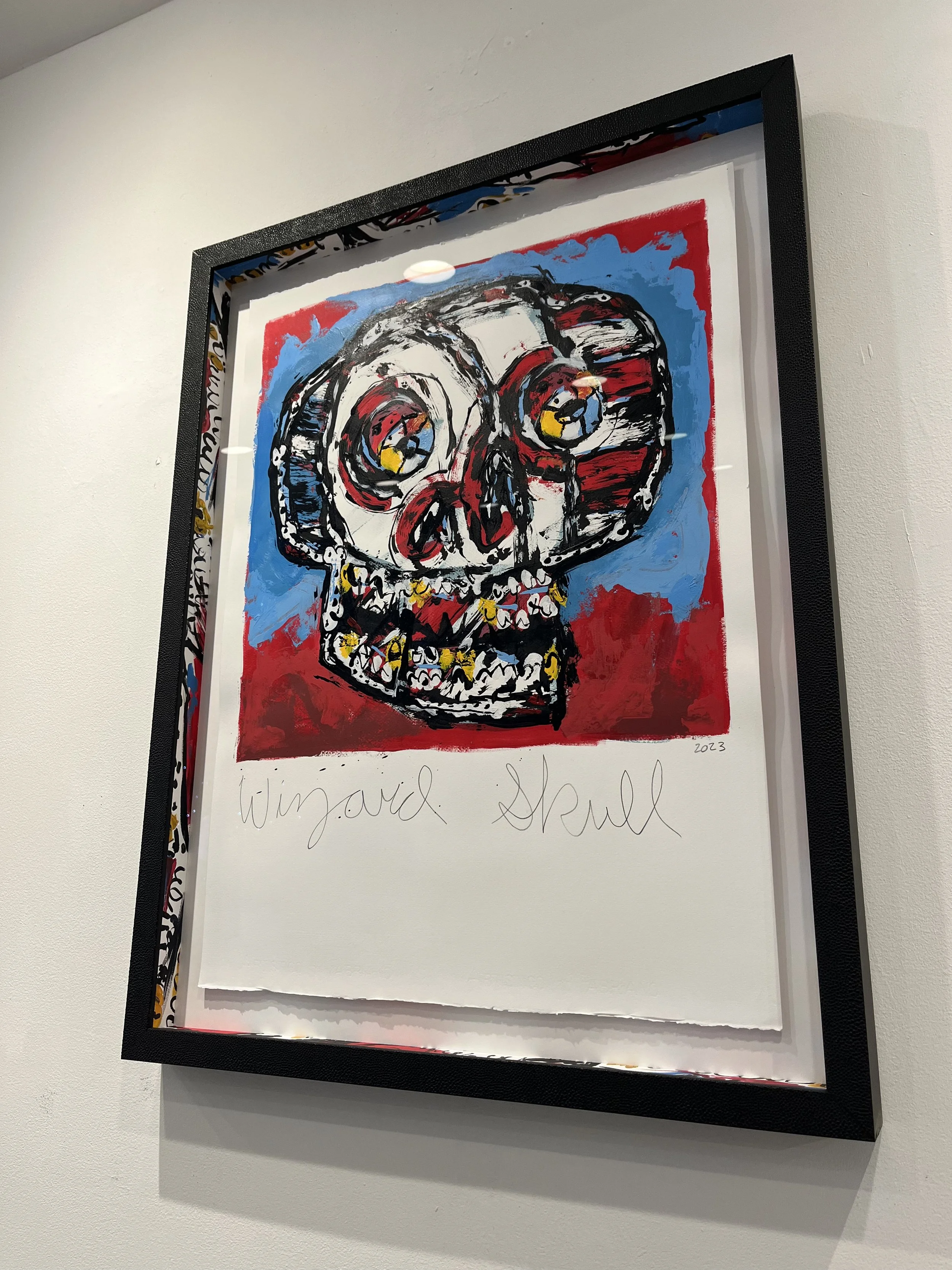 Colorful artwork of a skull with exaggerated features, painted with red, blue, yellow, and black. The piece is framed and includes the handwritten words "Wijdal Skull" and the year "2023" at the bottom.
