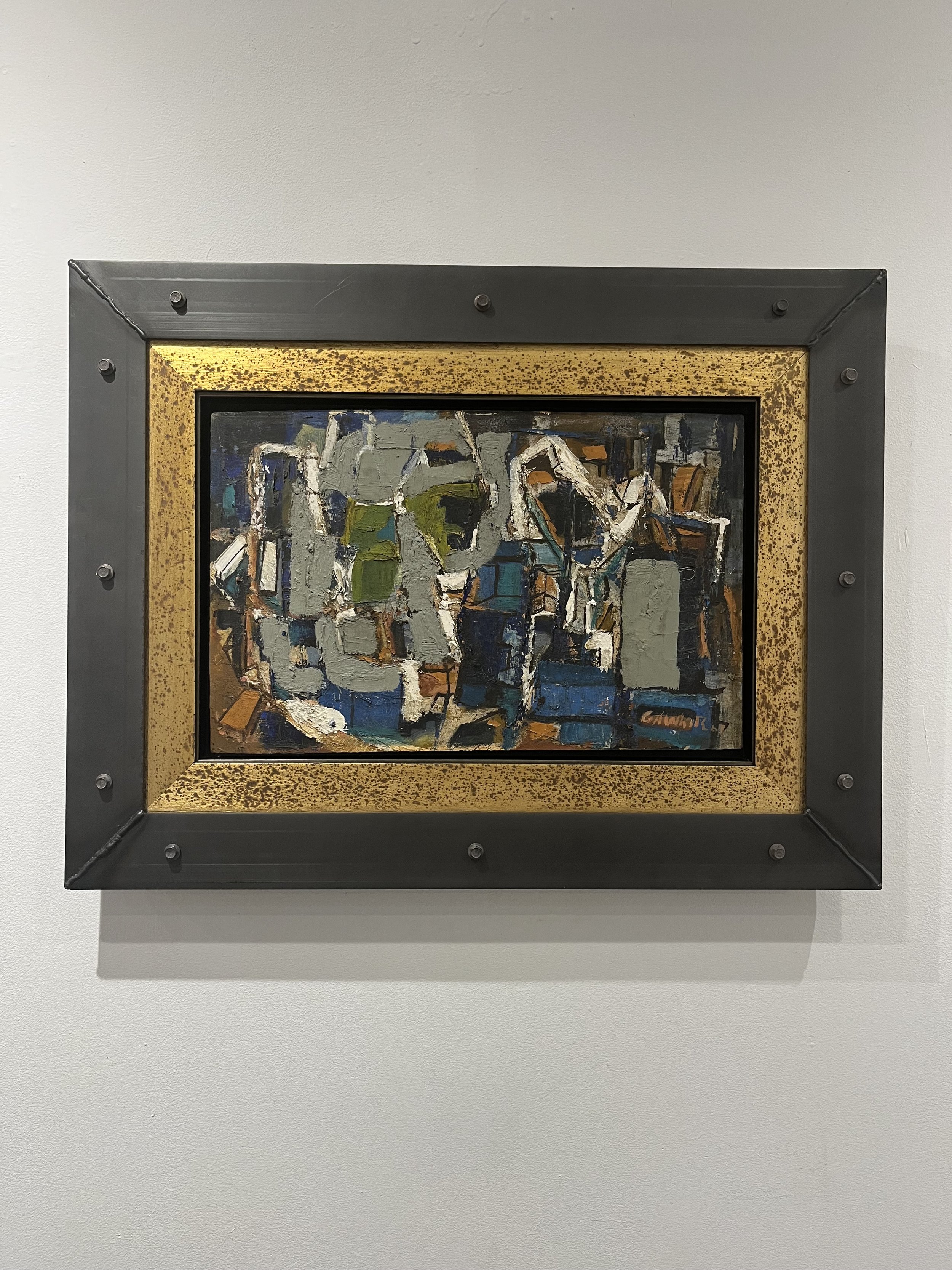 Abstract painting of a cityscape with buildings in shades of gray, green, blue, and brown, enclosed in a black and gold frame, displayed on a white gallery wall.
