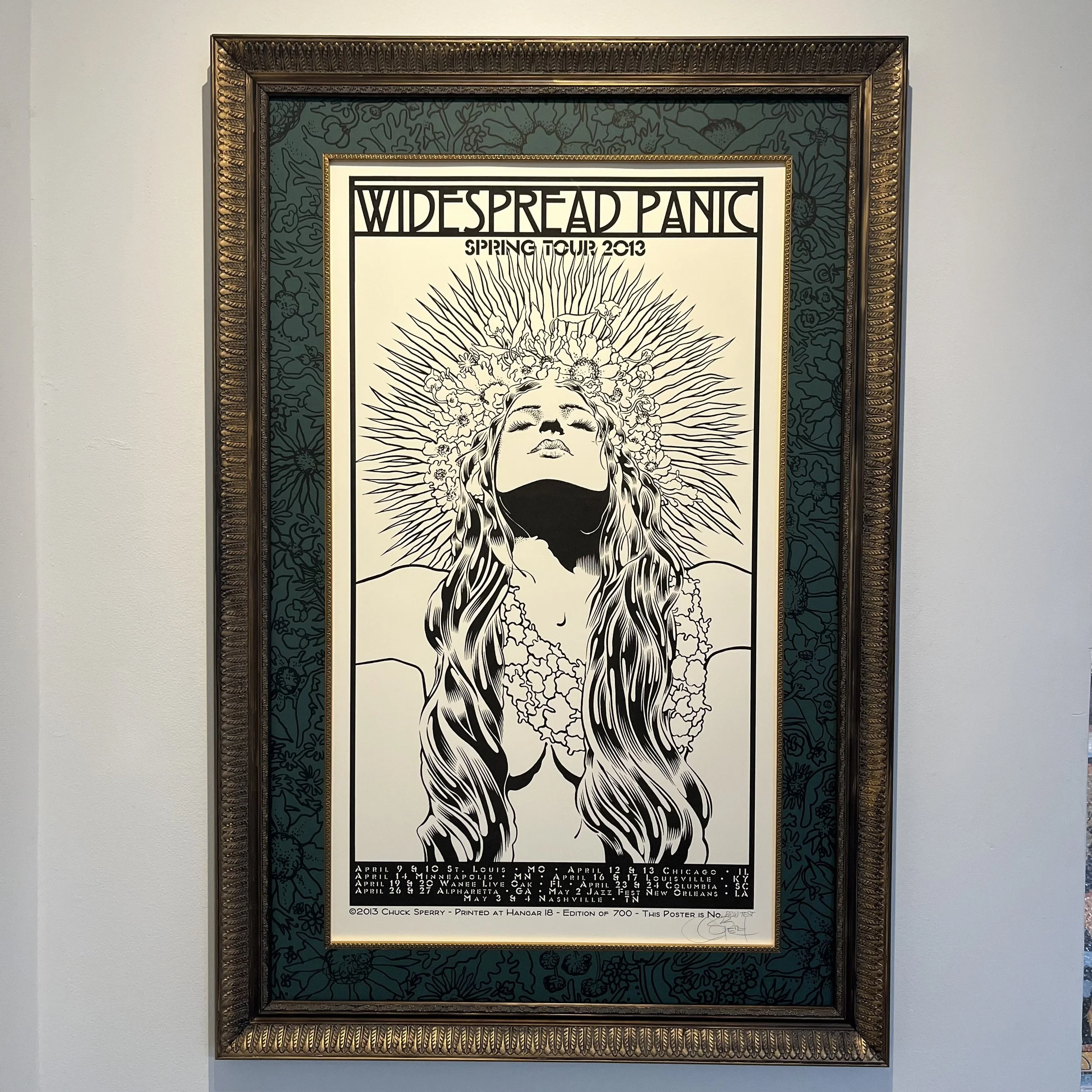 A framed poster for the band Widespread Panic's 2013 spring tour. The poster features a black and white illustration of a woman's face with long hair, floral elements, and radiating lines.