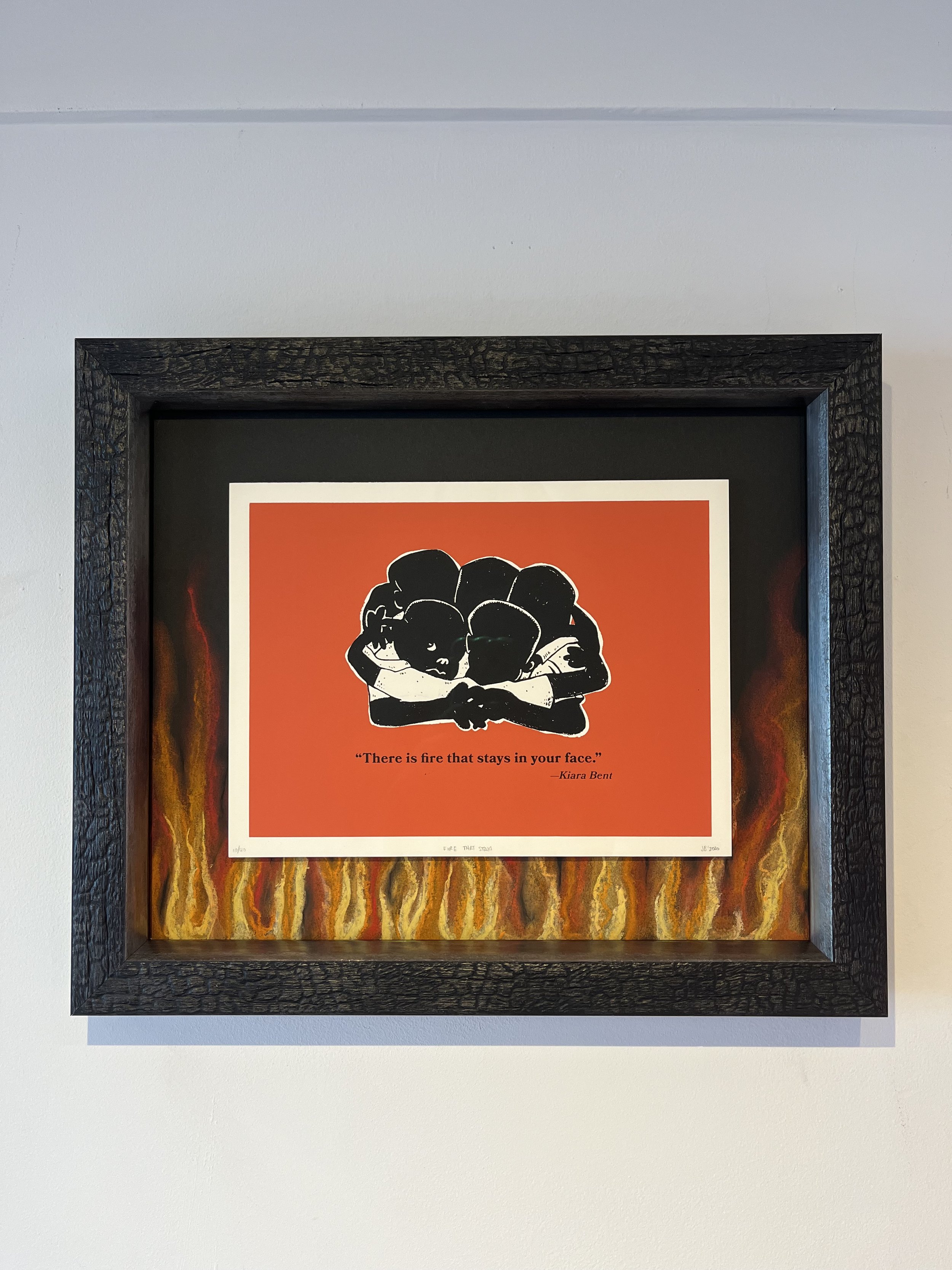 Artwork with a black and white illustration of four people embracing, set against an orange background, framed with a black wooden frame. Below the image, there is a quote: "There is fire that stays in your face." by Kiara Bent. The artwork is surrou
