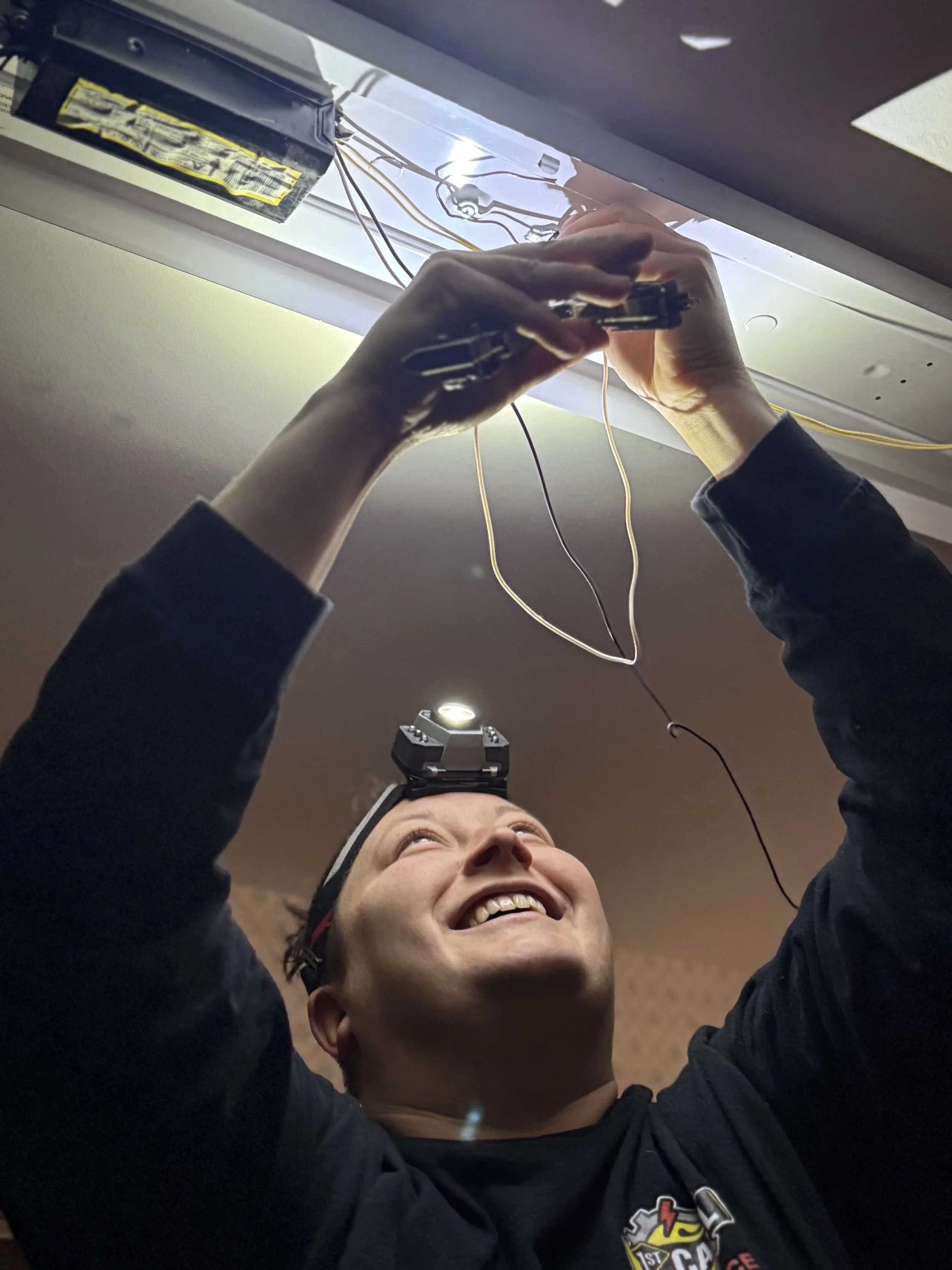 A person is installing or repairing an electrical fixture on the ceiling, with wires and a power supply visible. The person is smiling and wearing a black shirt with a logo.