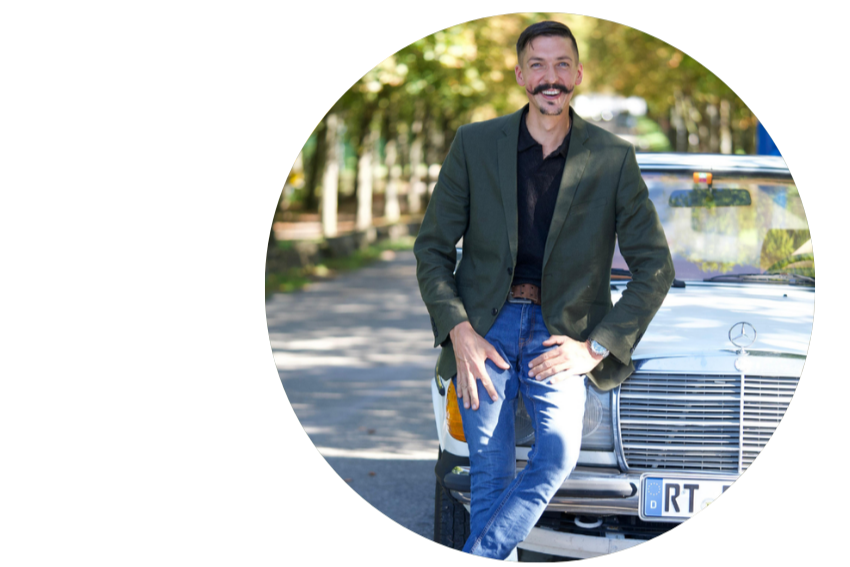 Ein Mann steht vor einem weißen Mercedes-Benz Auto auf einer Straße im Freien, umgeben von Bäumen, und lächelt in die Kamera.