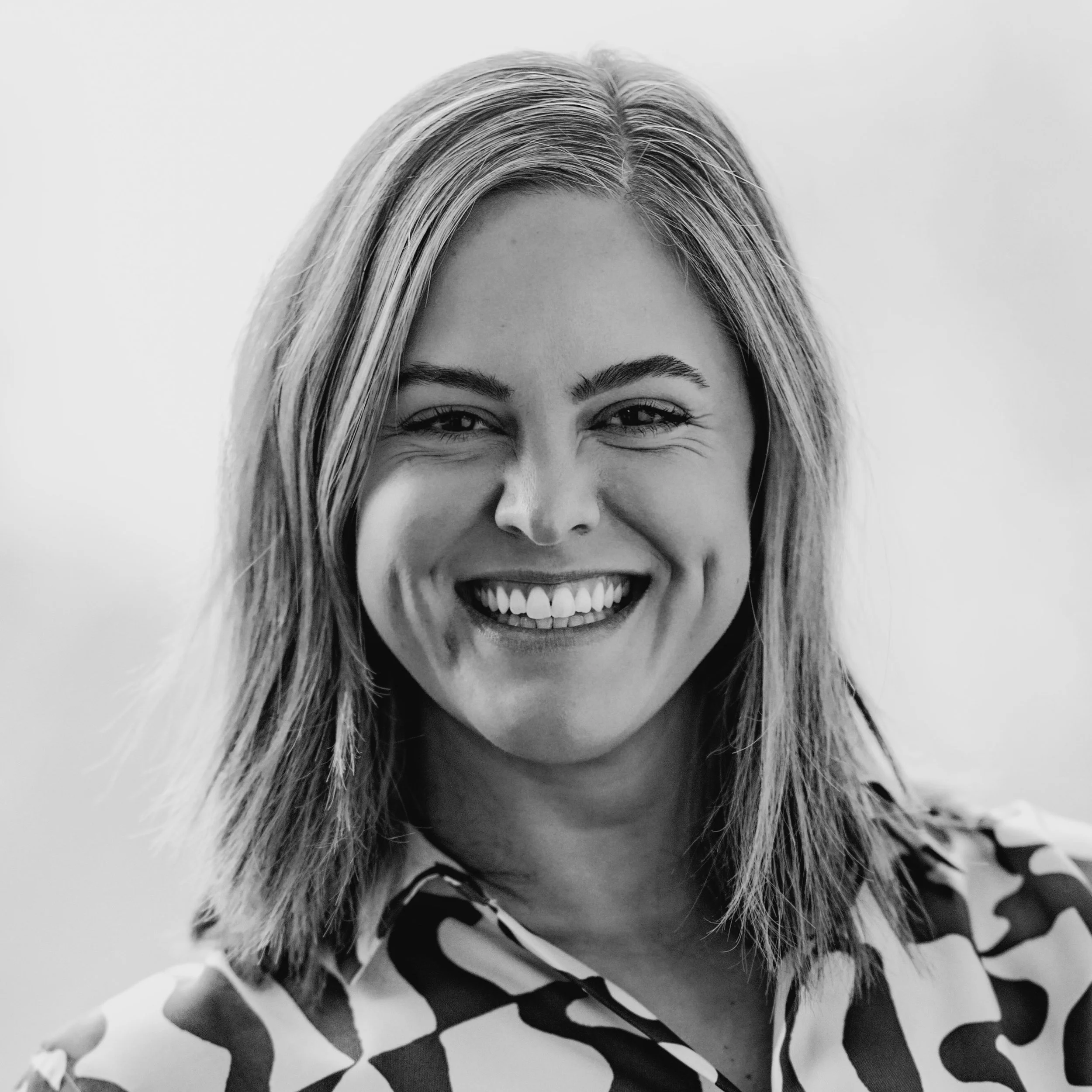 Black and white photo of Caitlin with light, straight hair smiling broadly, showing her teeth, with a smooth background.