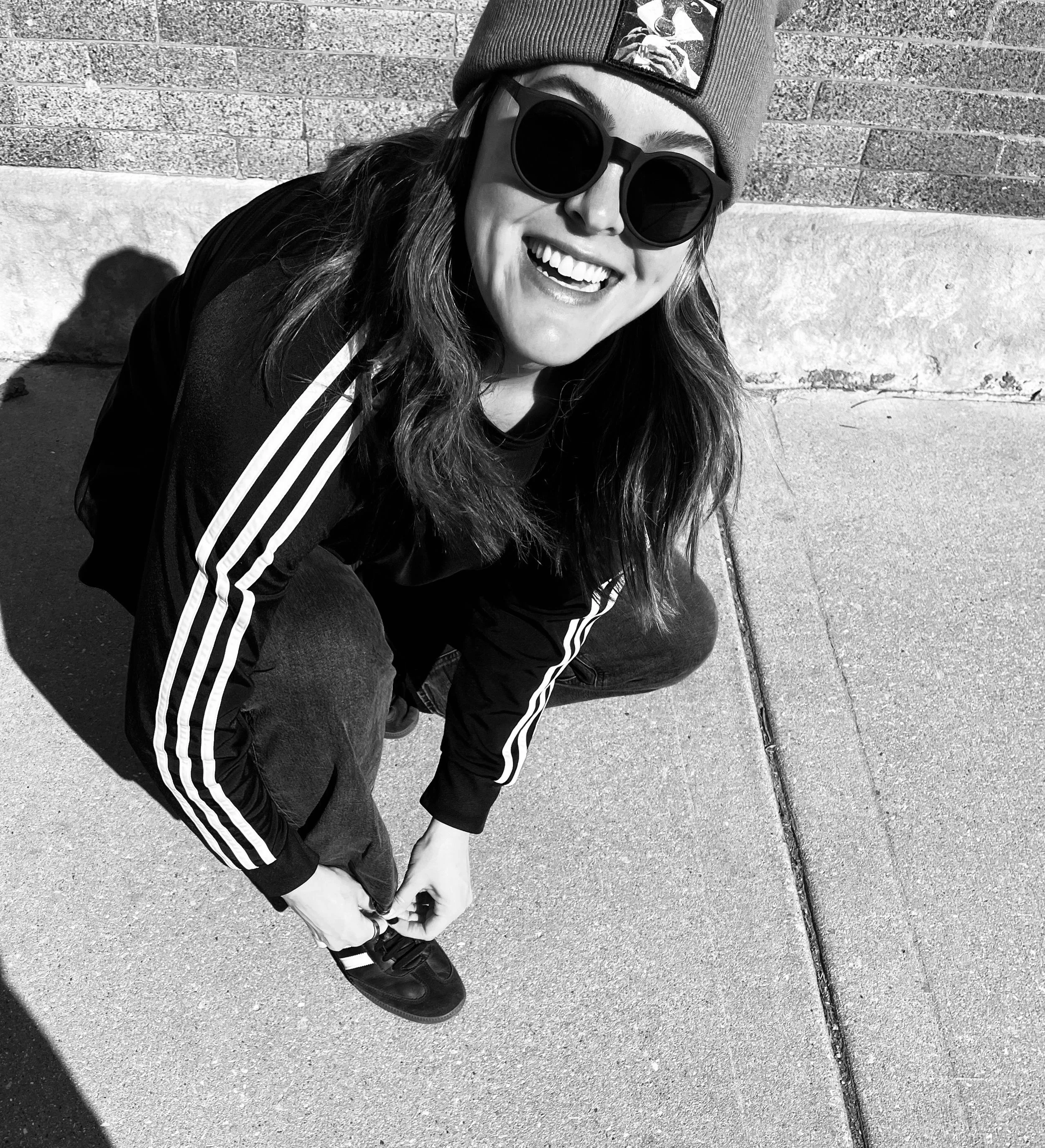 Caitlin smiling, wearing round sunglasses and a beanie hat with a patch, sitting on the ground against a brick wall background.