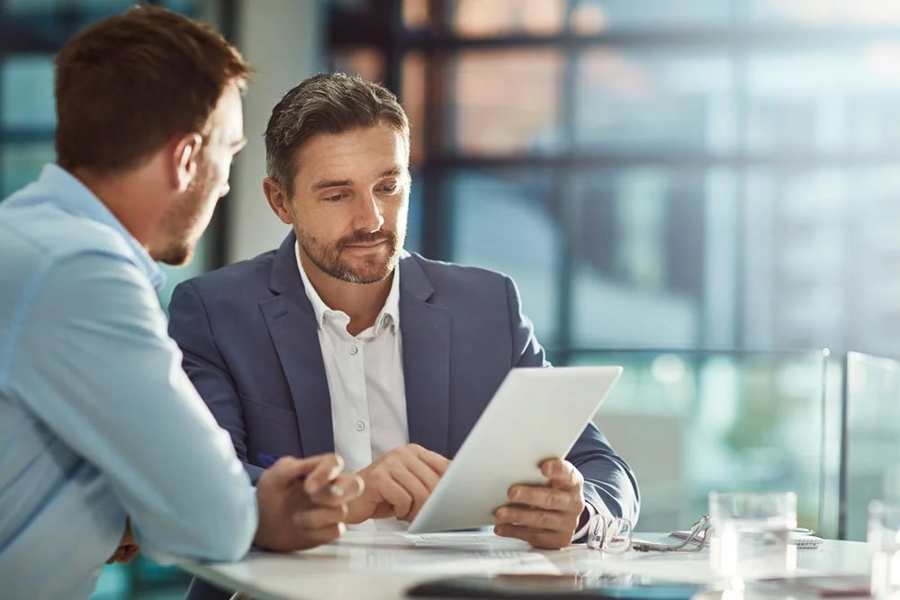 Two men in business attire sitting at a table in a modern office, looking at a tablet device.