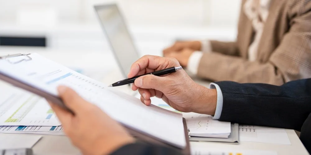 Close-up of a person's hand holding a pen and reviewing documents during a meeting, with another person working on a laptop in the background.