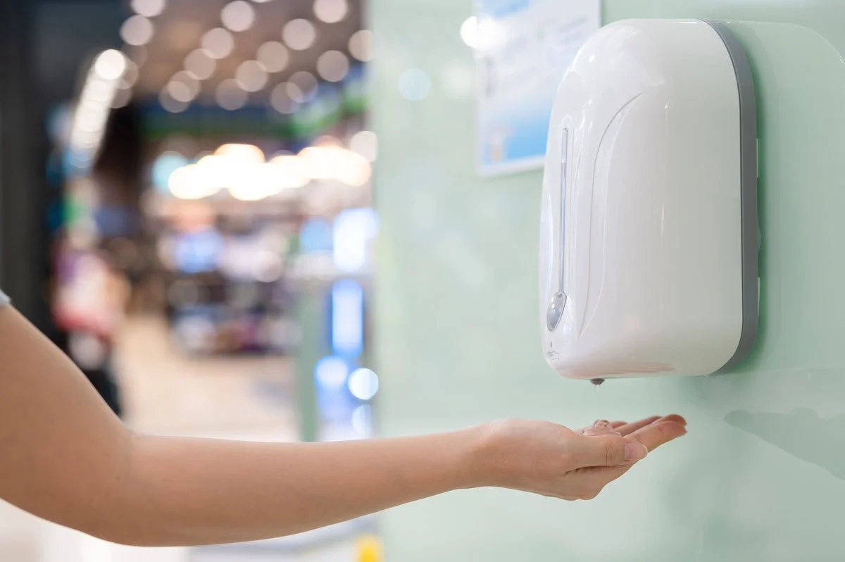 Person's hand under a wall-mounted automatic hand sanitizer dispenser in a public space.