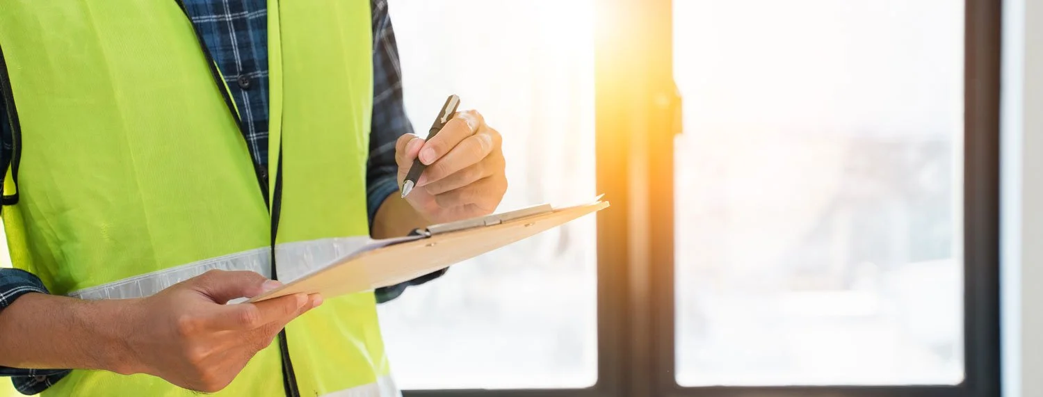 Person wearing a yellow reflective safety vest and a plaid shirt, writing on a clipboard with a pen near a window with sunlight.