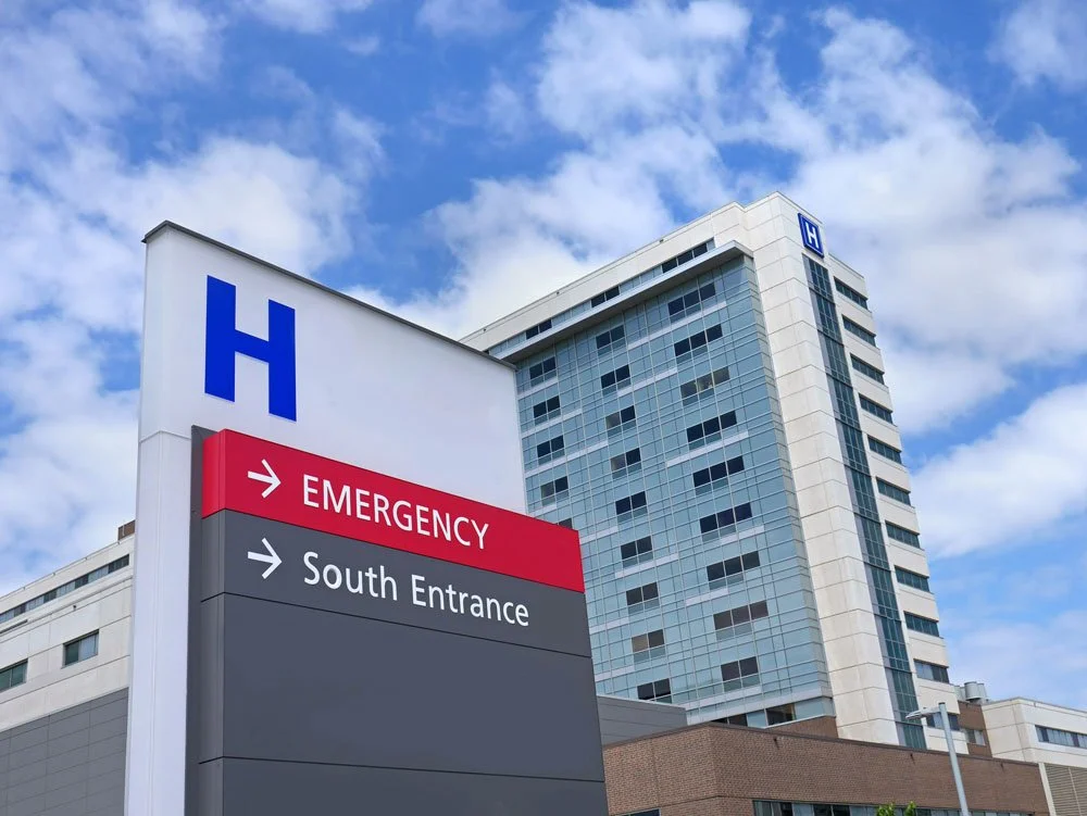 Hospital emergency sign indicating the emergency entrance and south entrance, with a high-rise hospital building in the background under a partly cloudy sky.