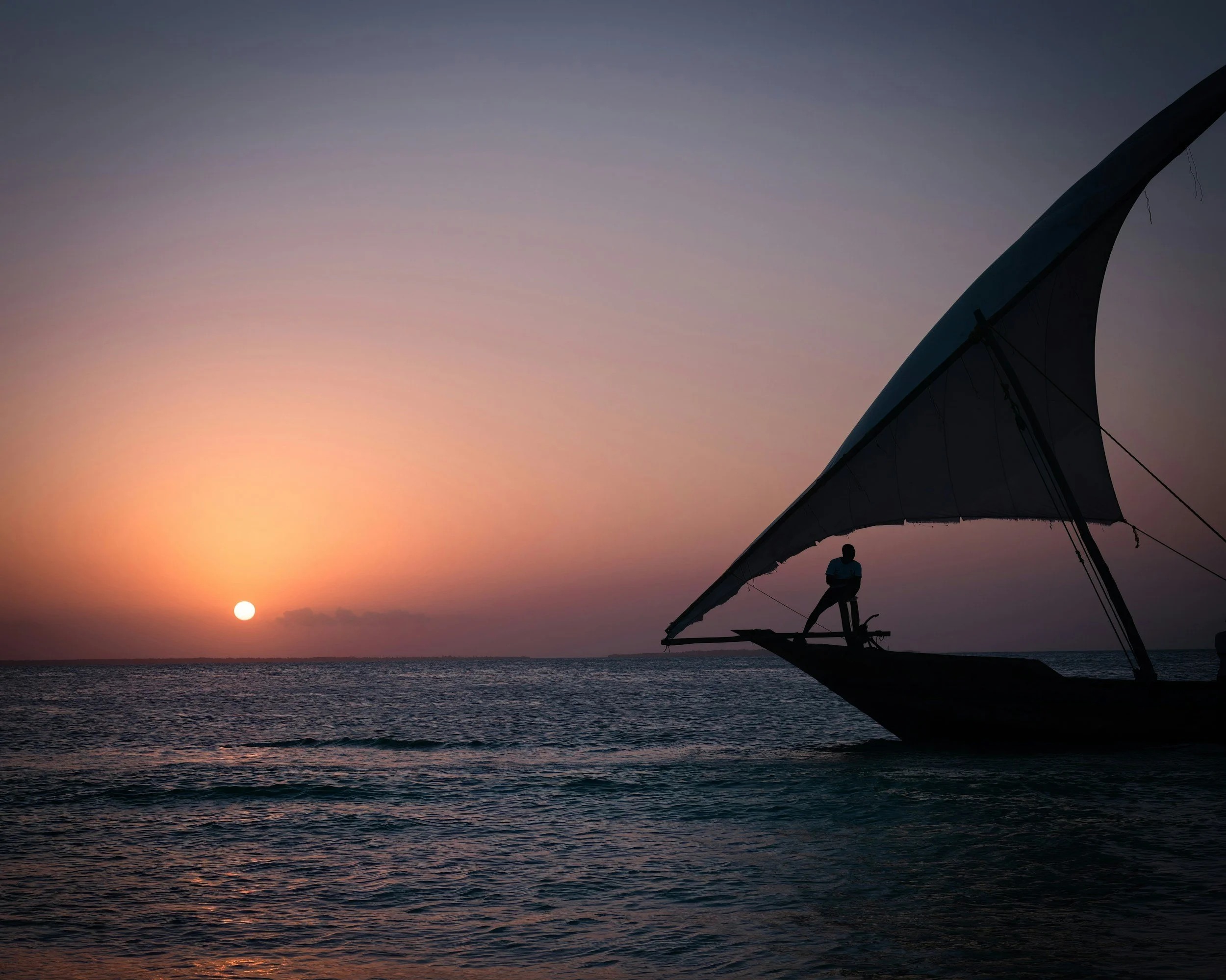 Silhouette of a person standing on the bow of a traditional fishing boat with a large sail, during a sunset over calm ocean waters.