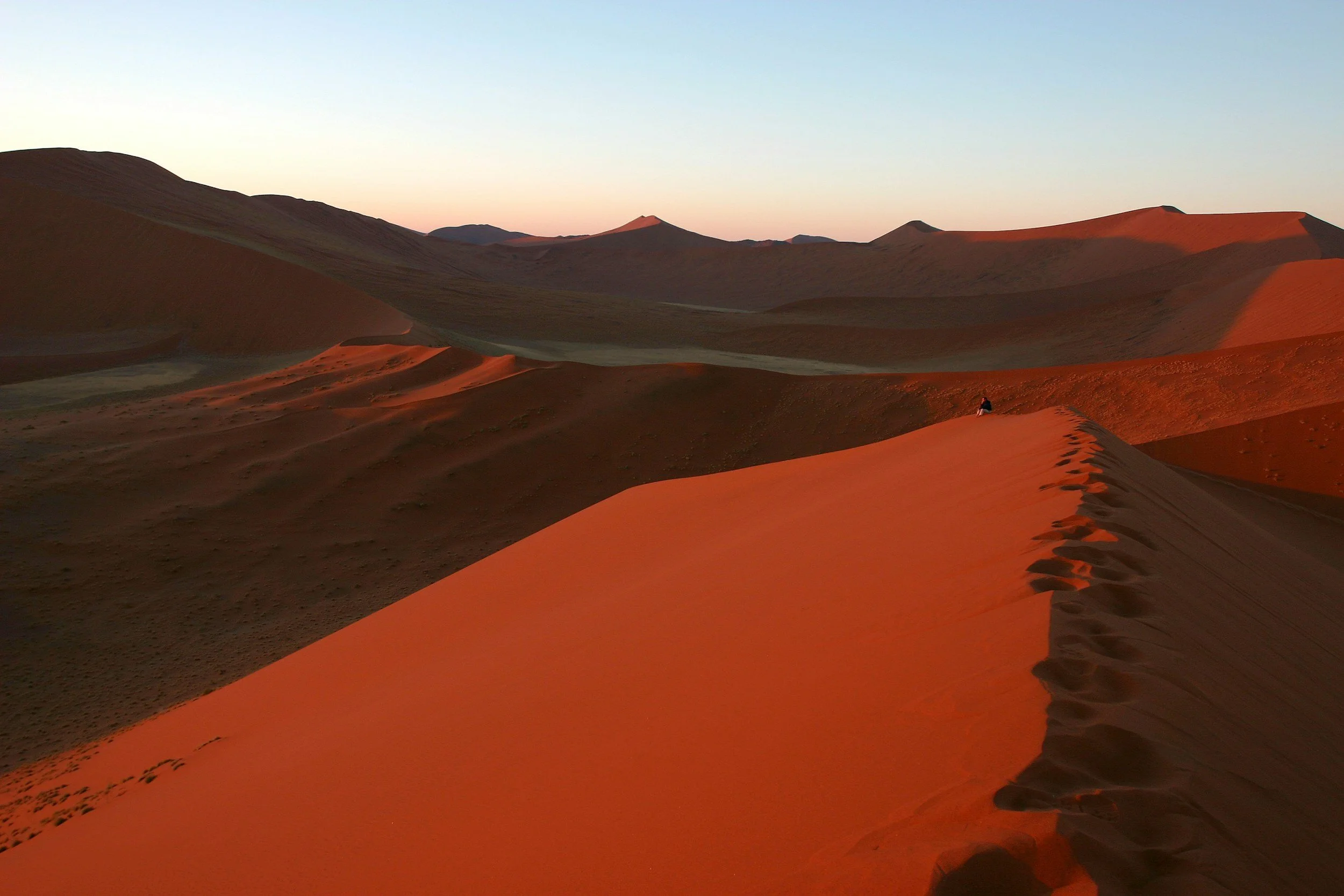 Desert landscape with orange sand dunes, some people hiking along a ridge, and a clear sky during sunset or sunrise