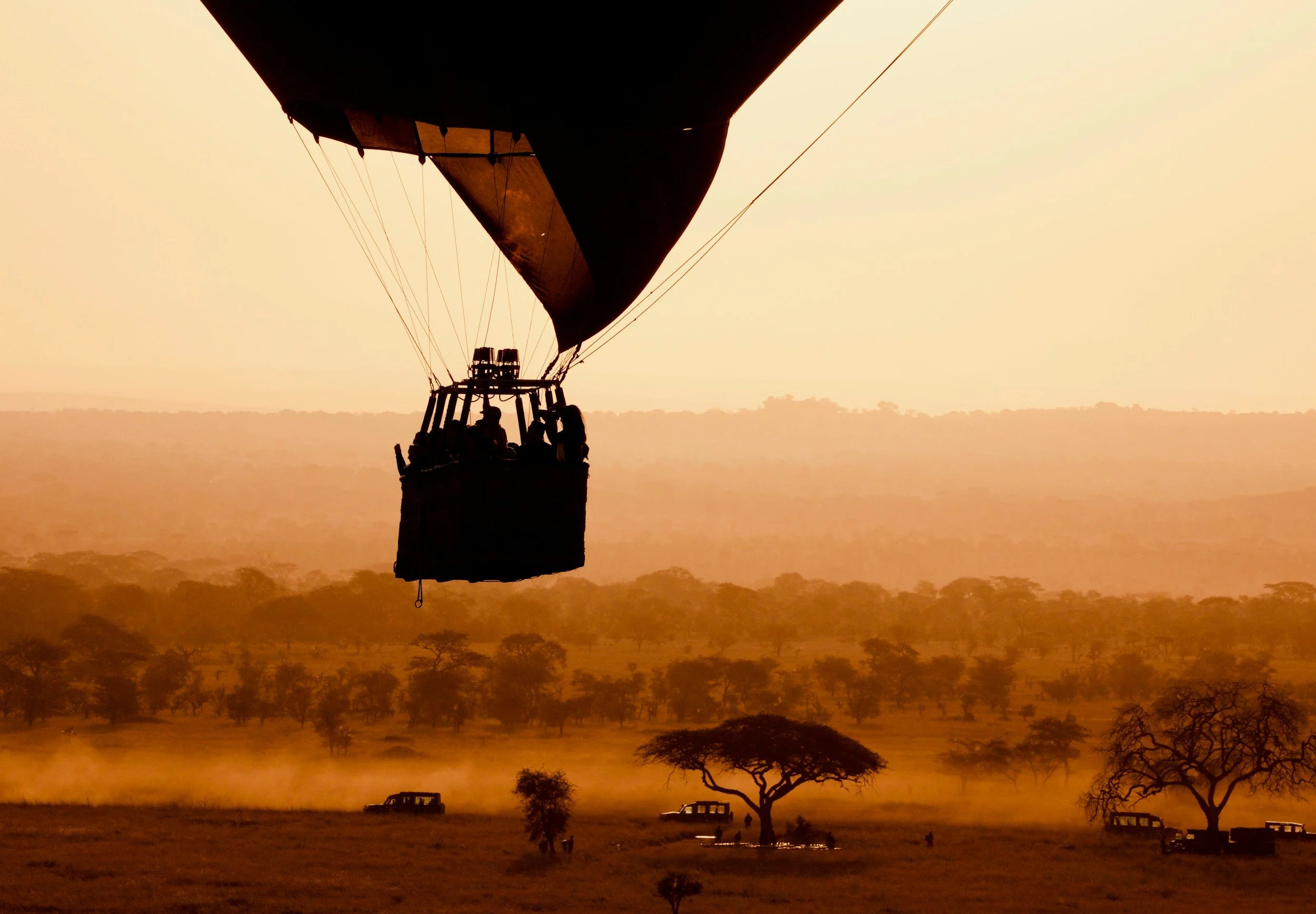 Hot air balloon flying over a savannah landscape with sparse trees and several safari vehicles at sunset.
