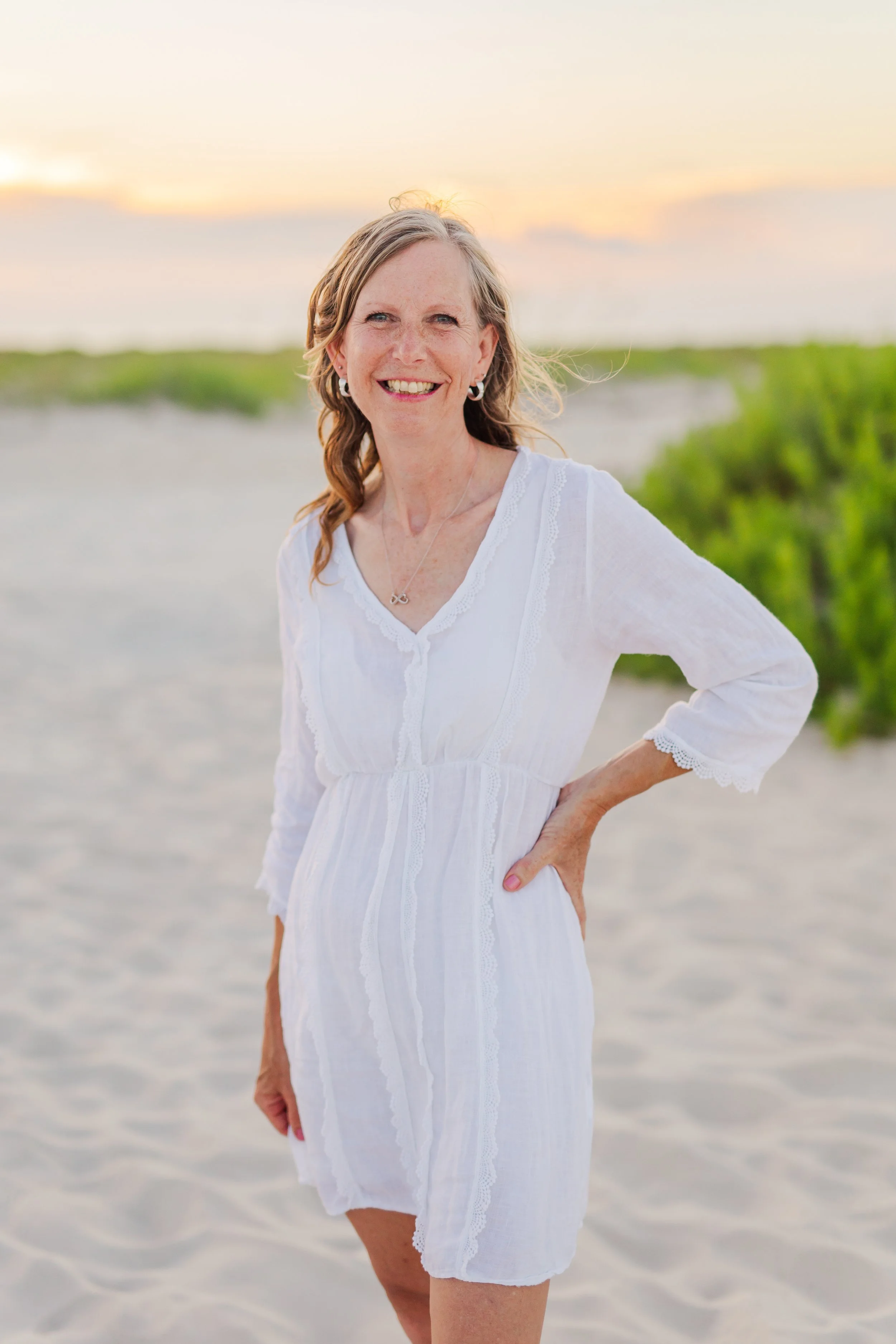 Smiling woman in a white dress standing on sandy beach during sunset, with greenery and sky in the background.